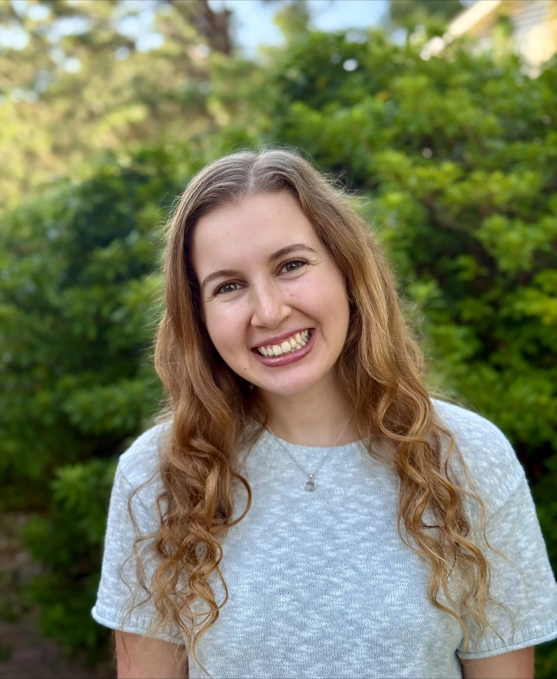 Somdal Associates architecture interior design, louisiana, shreveport, baton rouge, Young woman with long, wavy brown hair smiling outdoors in front of green bushes and trees.