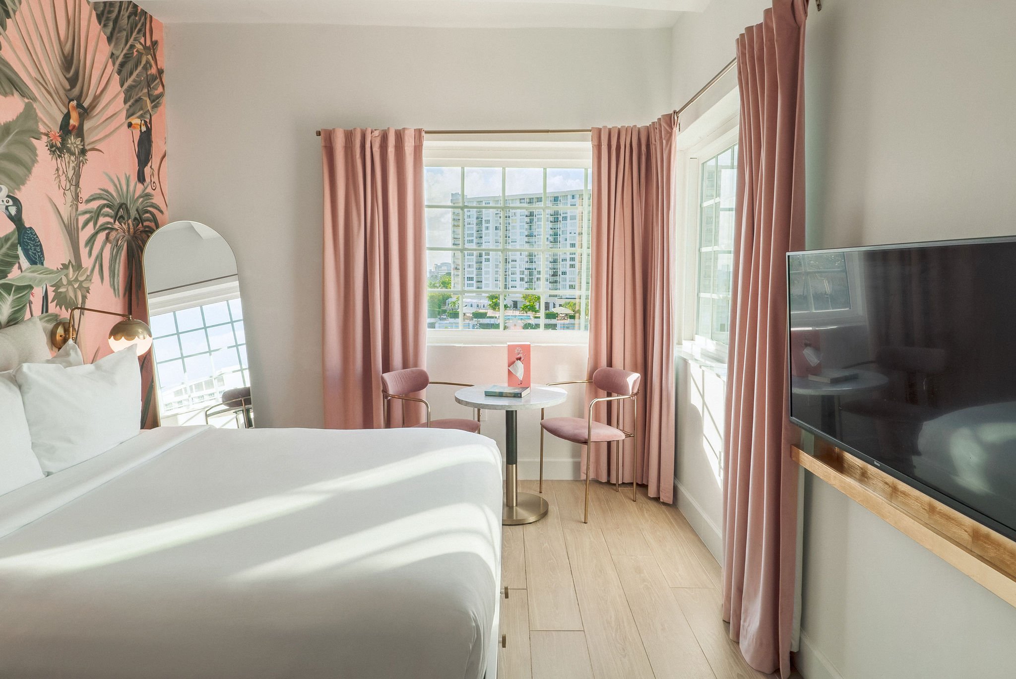 Bright boutique hotel room at Le Particulier Miami featuring a king bed with plush pillows, pastel pink curtains, tropical mural, gold lighting, modern chandelier, flat screen TV, and a cozy seating area by large corner windows with water views