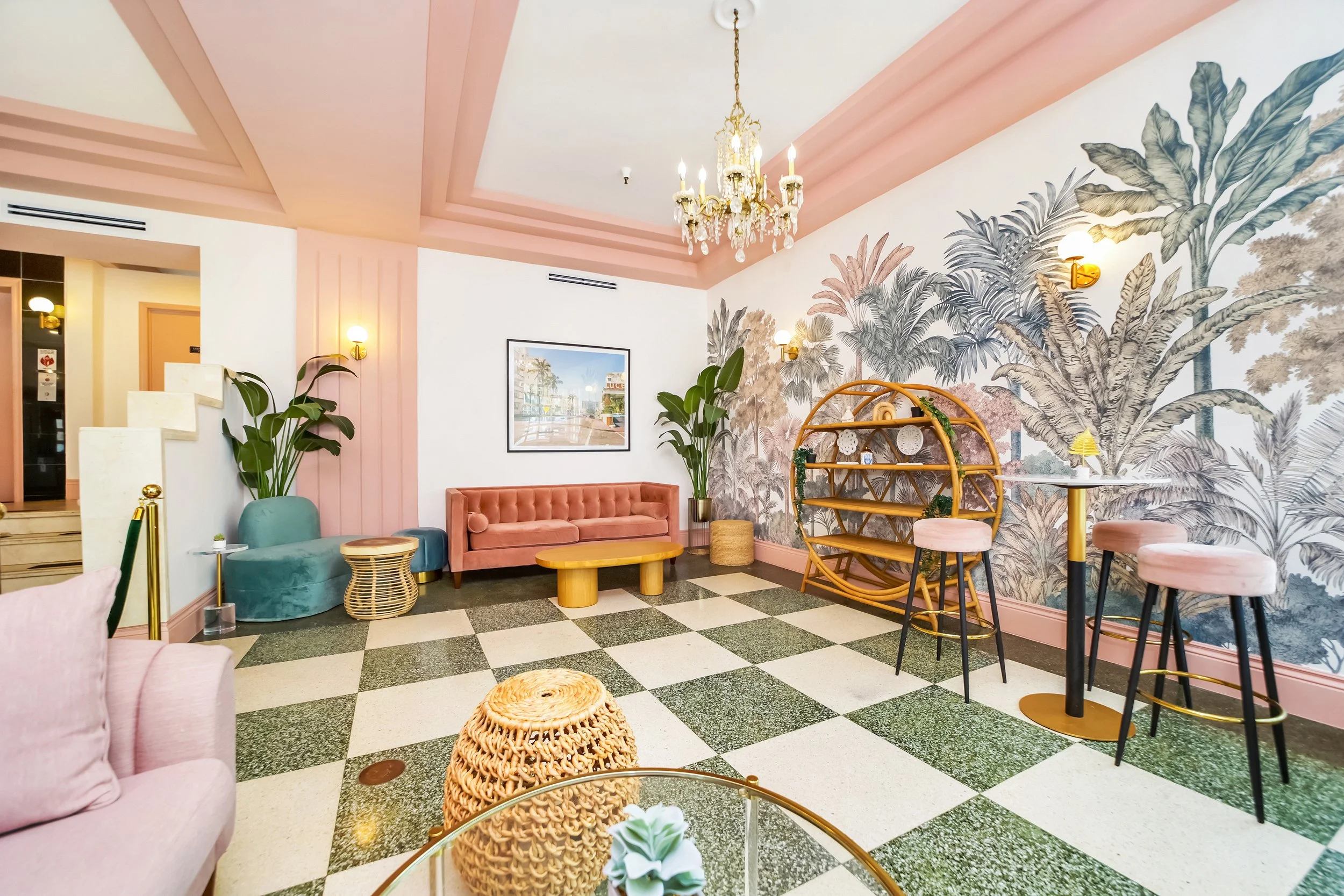 Chic lobby at Le Particulier Miami featuring a pastel pink velvet sofa, tropical botanical wallpaper, gold chandelier, rattan furniture, and checkered tile floor