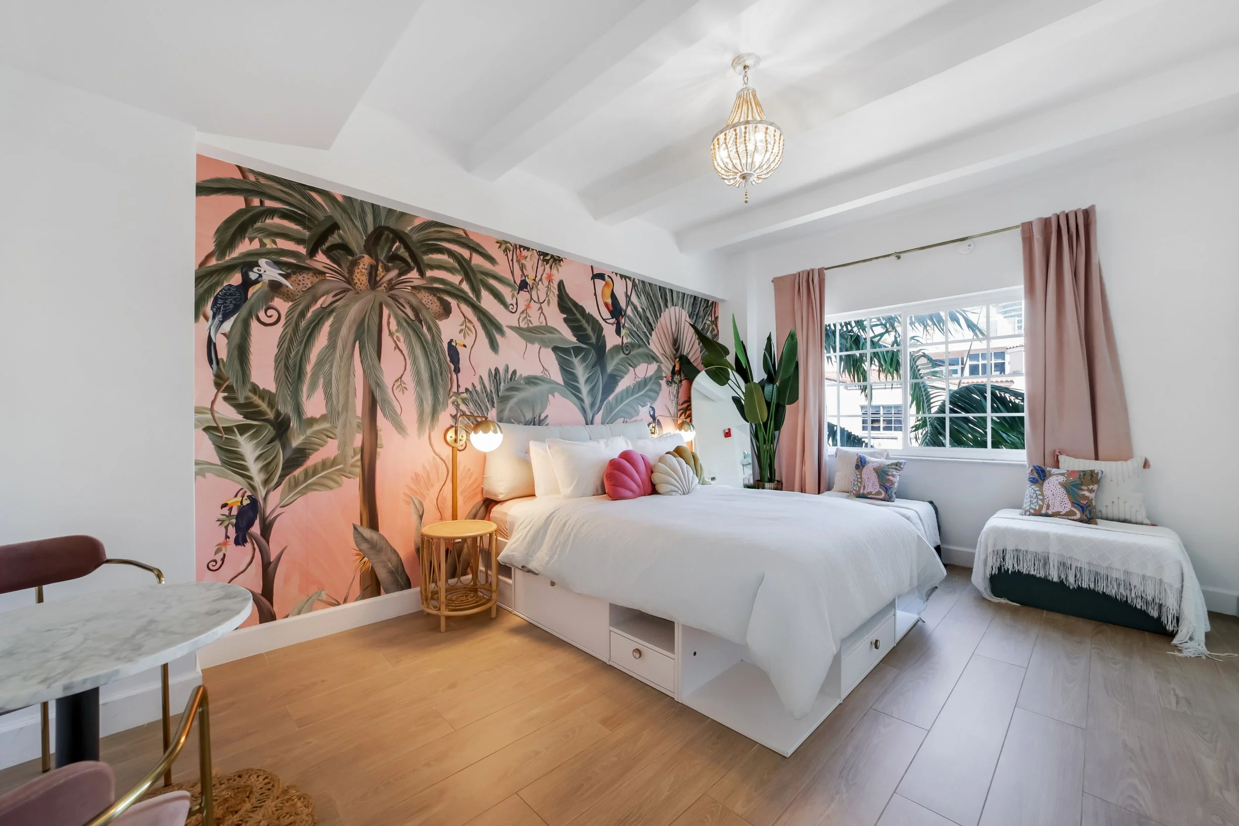 Bright boutique hotel room at Le Particulier Miami featuring a king bed with storage, tropical mural with toucans and palm leaves, pastel pink curtains, cozy daybed by a large window with city and ocean views, modern gold lighting, and chic decor
