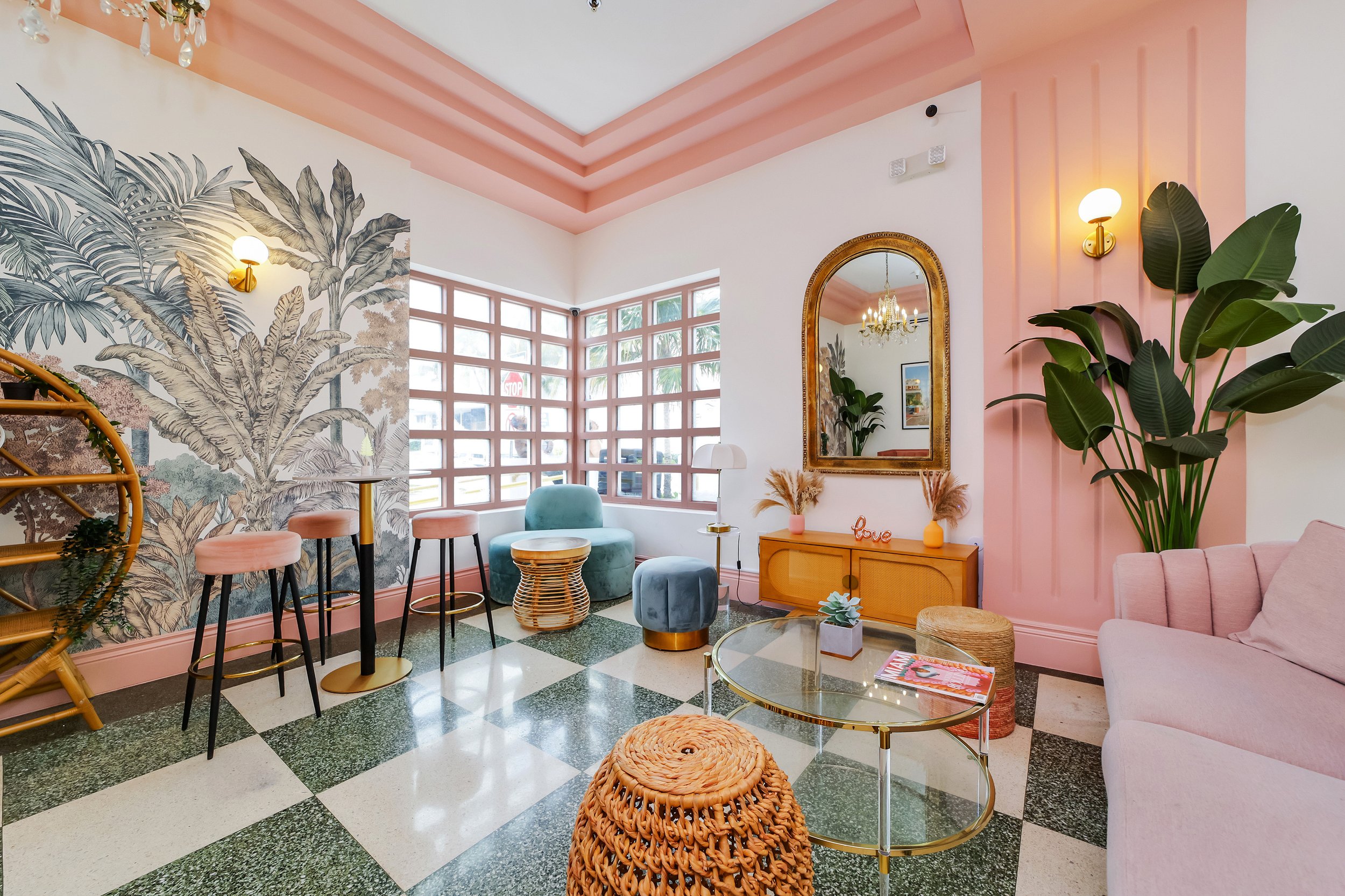 Bright and stylish lounge area at Le Particulier Miami featuring pastel pink walls, checkered tile floors, rattan stools, modern velvet seating, gold accents, a large mirror, and tropical decor