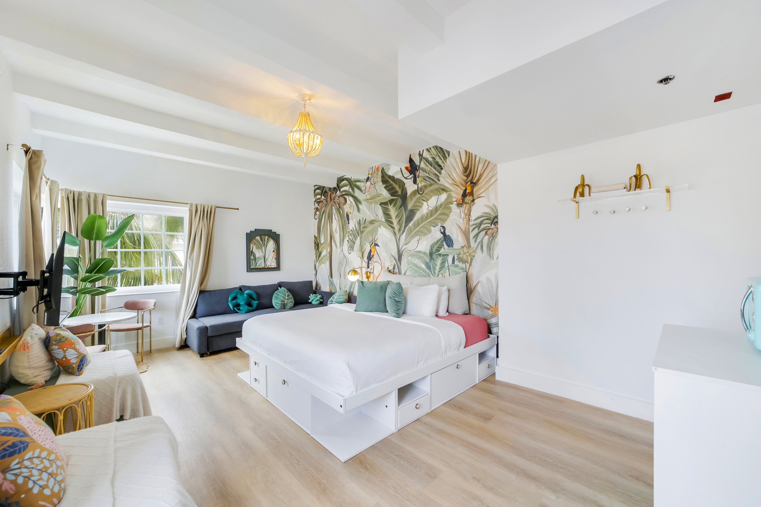 Spacious boutique hotel room at Le Particulier Miami featuring a king bed with storage drawers, a tropical mural with toucans and palm leaves, modern sofa, natural light from large windows, and chic decor