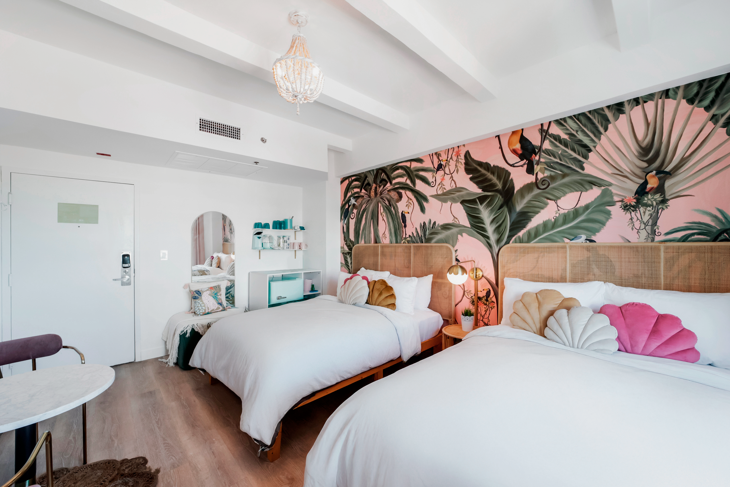 Bright boutique hotel room at Le Particulier Miami featuring two double beds with rattan headboards, shell-shaped velvet pillows, a tropical mural with toucans and palm leaves, pastel decor, and modern amenities