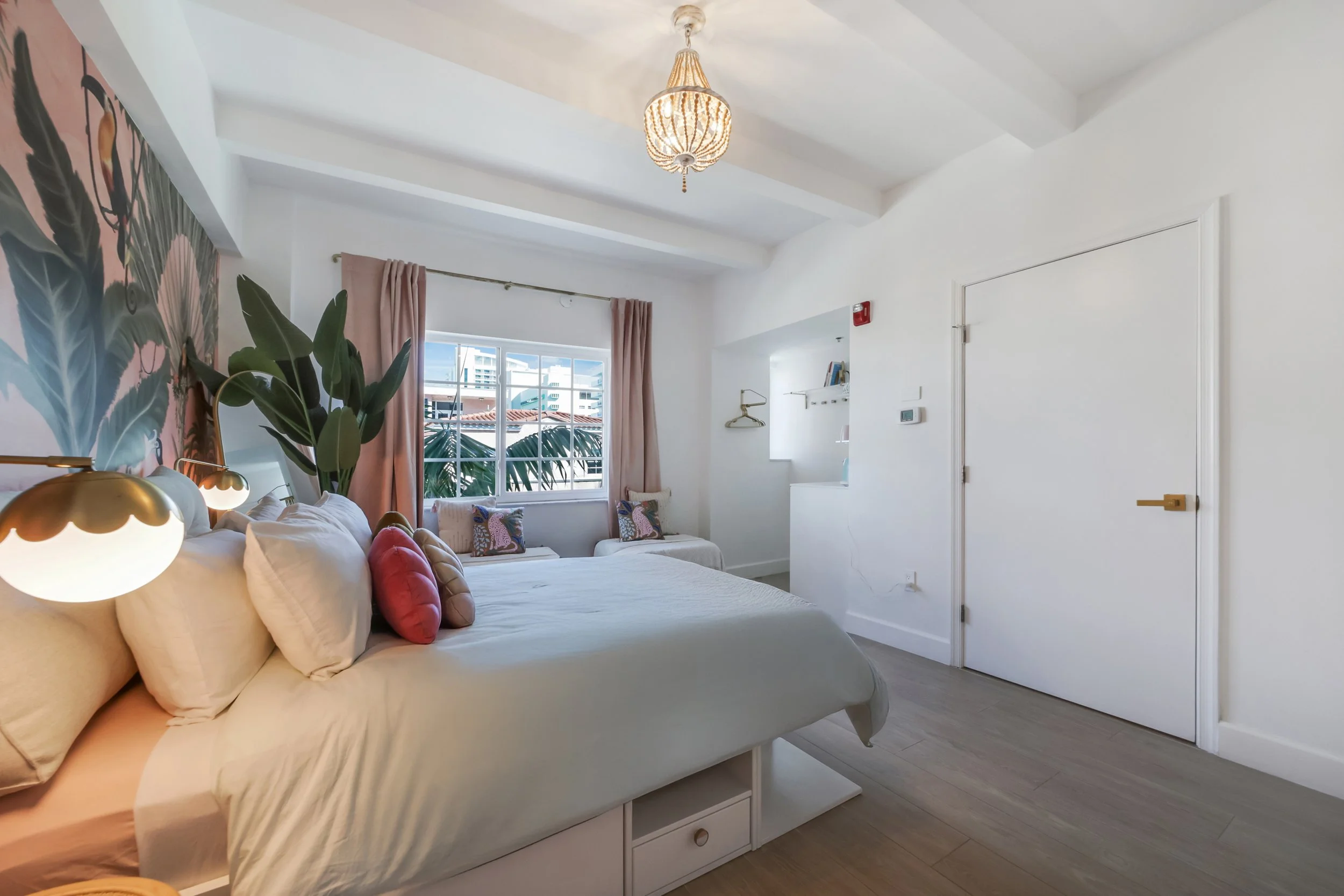 Bright and stylish boutique hotel room at Le Particulier Miami featuring a tropical mural, plush bedding, pastel pink curtains, modern gold lighting, and a cozy seating nook by large windows with city views.