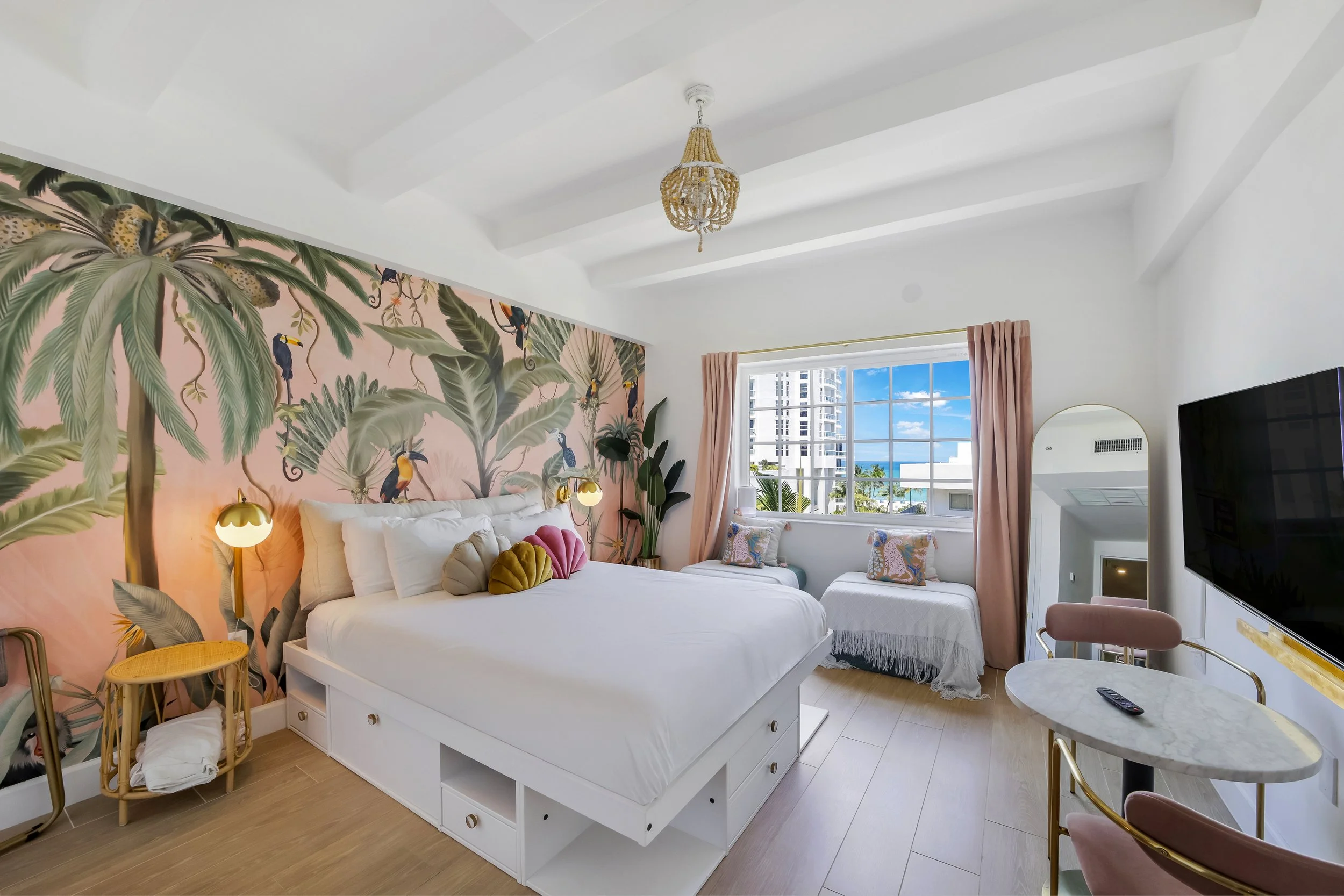 Bright boutique hotel room at Le Particulier Miami featuring a king bed with storage, tropical mural with toucans and palm leaves, pastel pink curtains, cozy daybed by a large window with city and ocean views, modern gold lighting, and chic decor