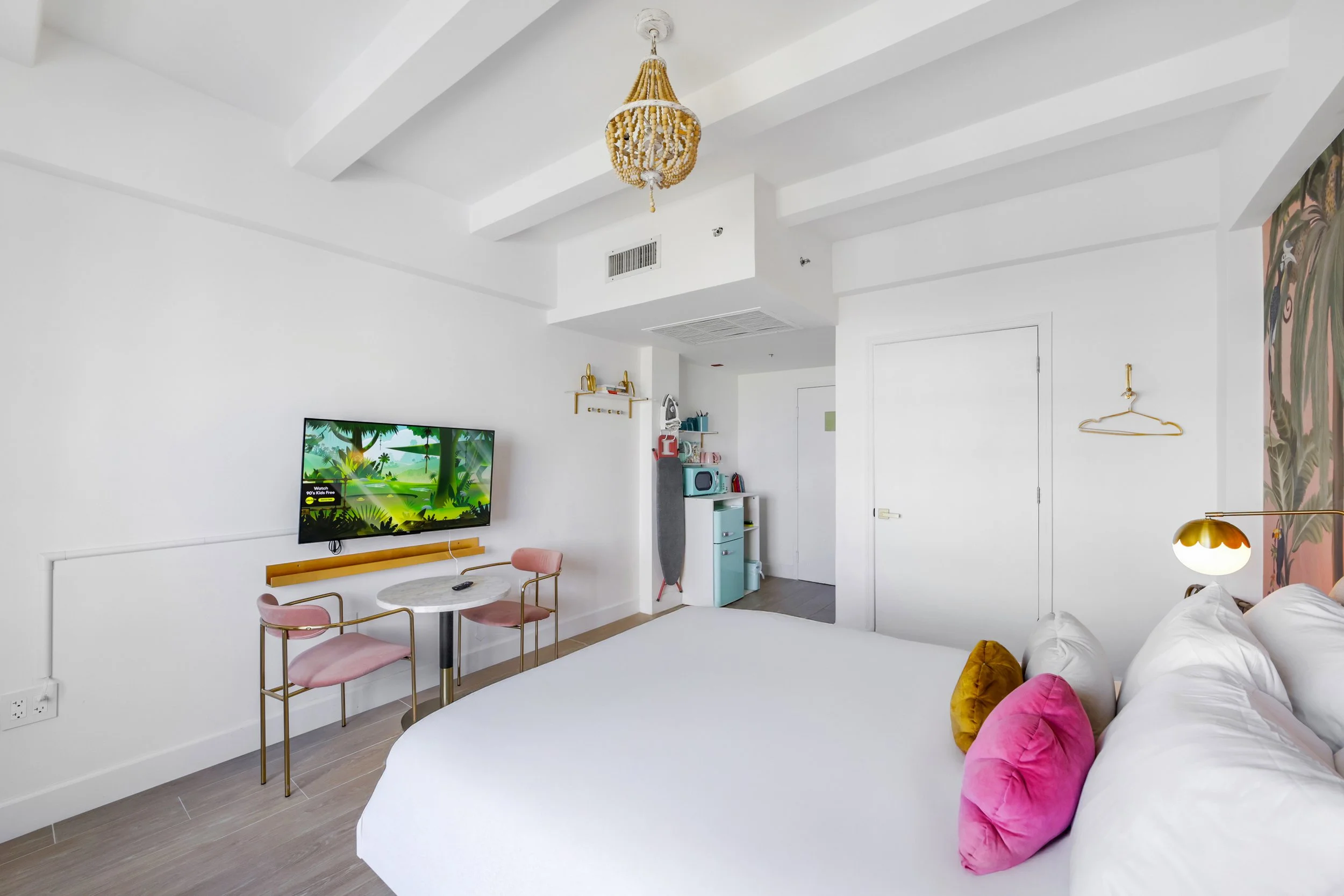 Bright and stylish boutique hotel room at Le Particulier Miami featuring a tropical mural, plush bedding, pastel pink curtains, modern gold lighting, and a cozy seating nook by large windows with city views