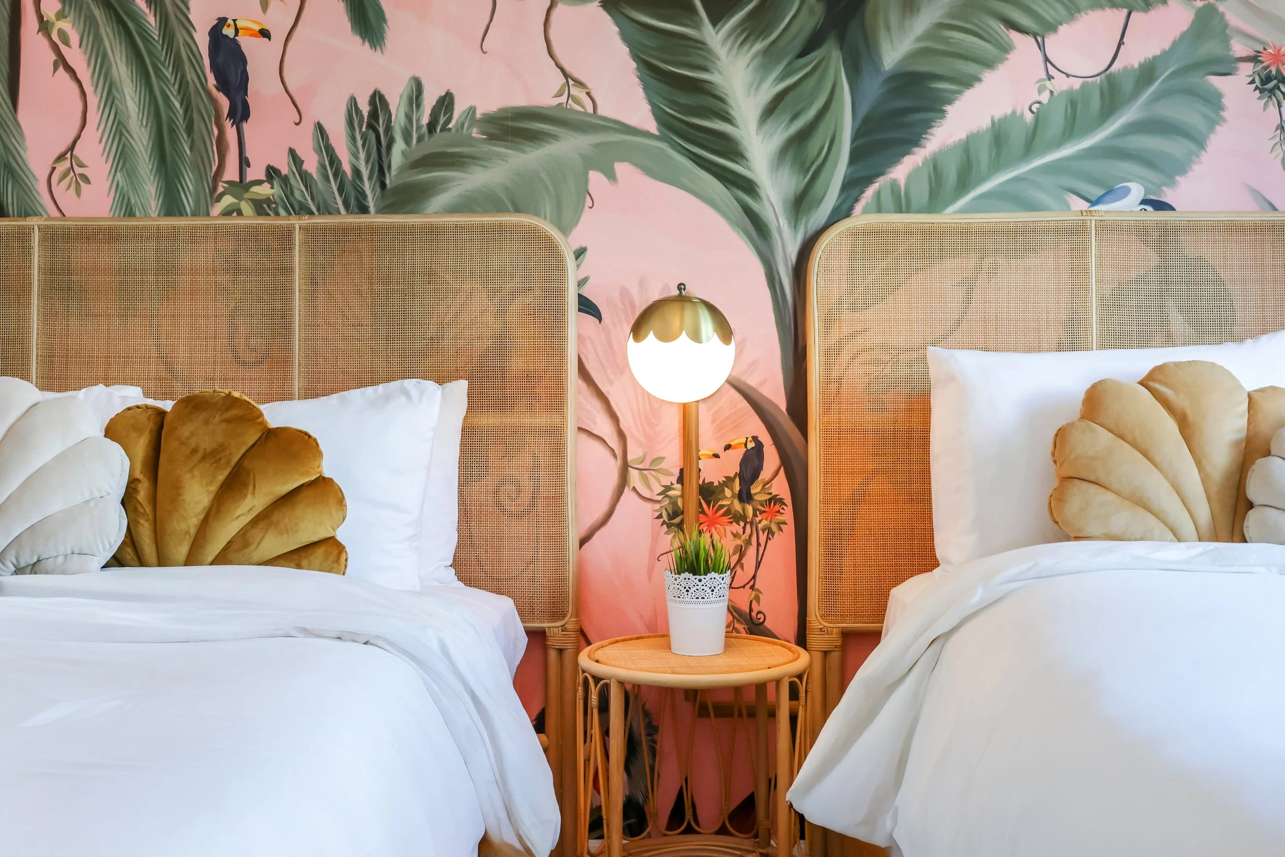 Two twin beds with rattan headboards, shell-shaped velvet pillows, and crisp white bedding, set against a tropical mural with toucans and palm leaves at Le Particulier Miami