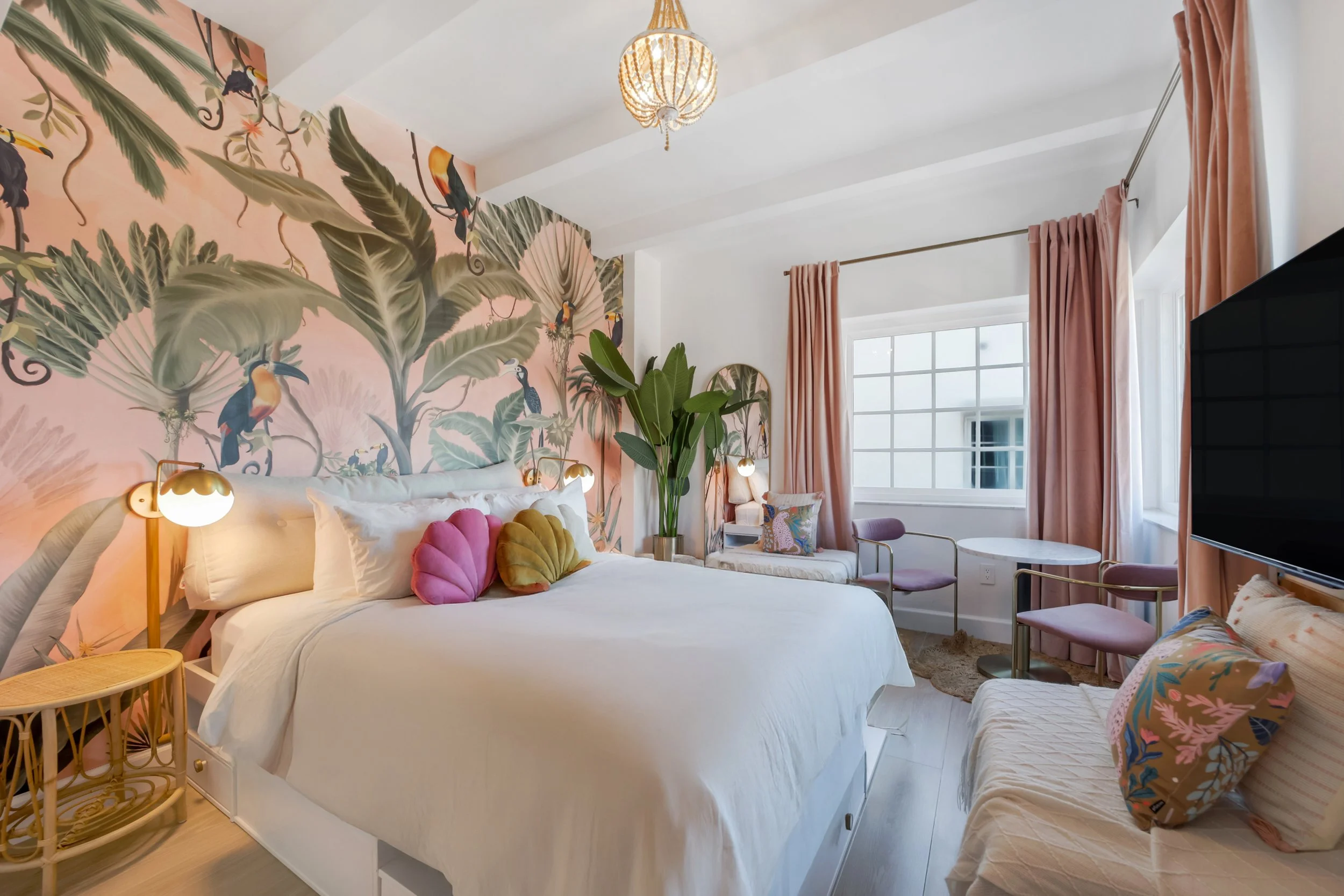 Boutique hotel room at Le Particulier Miami featuring a tropical mural with toucans and palm leaves, pastel pink curtains, plush bedding, and stylish decor