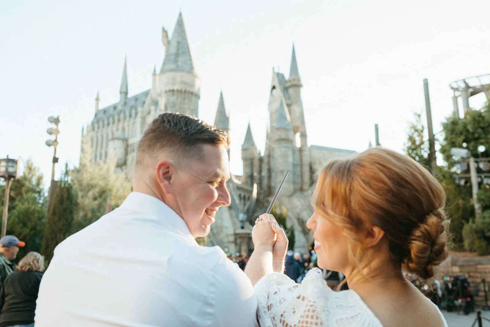 A man and woman smiling at each other in front of a castle, with the woman holding a wand, during an outdoor event at a theme park.