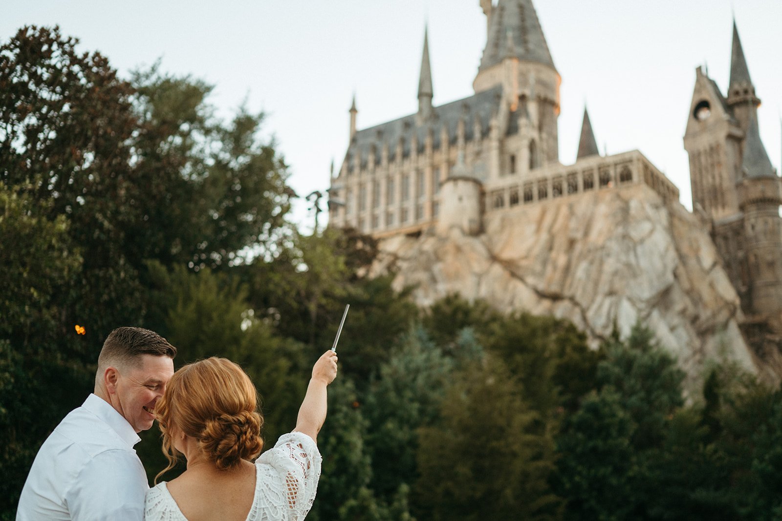A couple taking a selfie with a castle in the background, surrounded by trees.