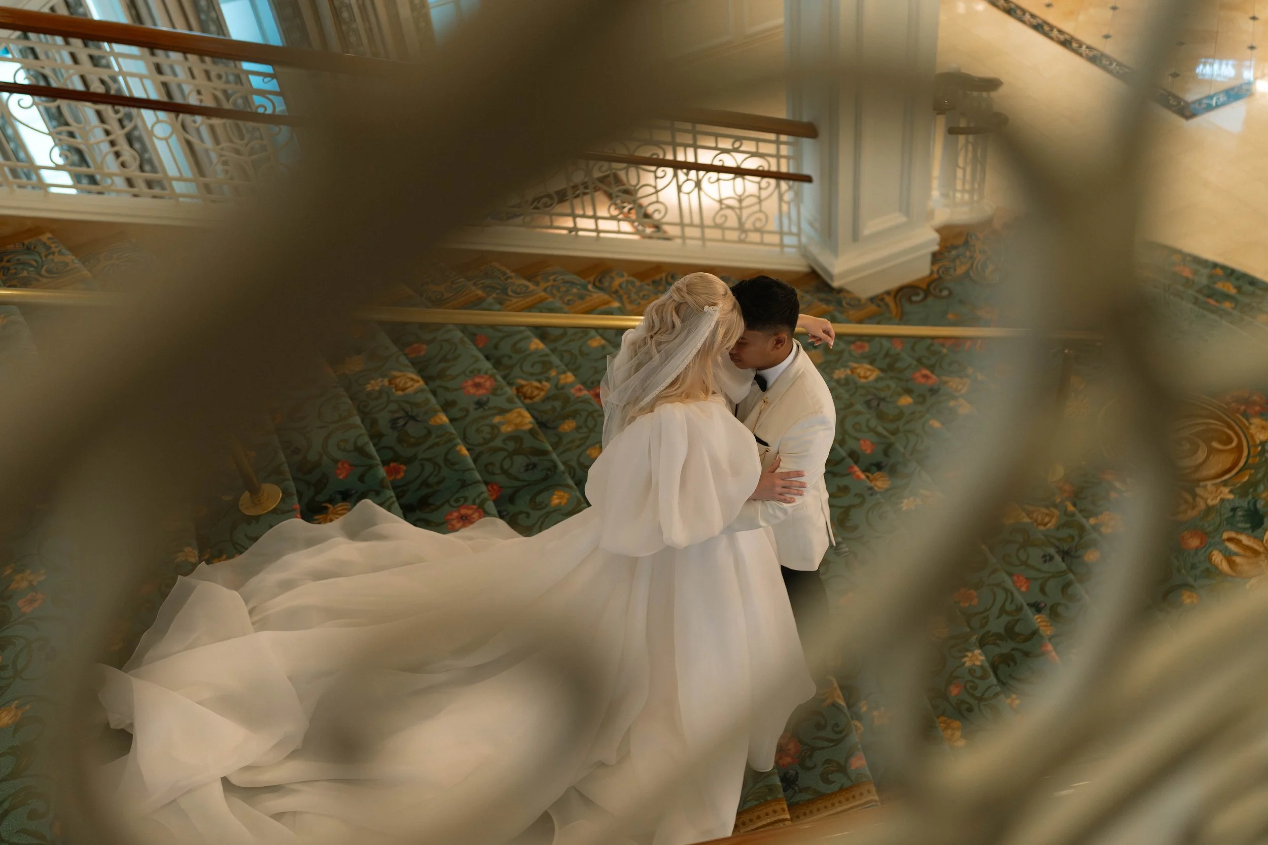 A bride and groom share a kiss on a staircase, viewed from above through a decorative railing. The bride wears a white gown with a long train, and the groom is in a white tuxedo with a black bow tie. The staircase has a floral-patterned carpet, and t