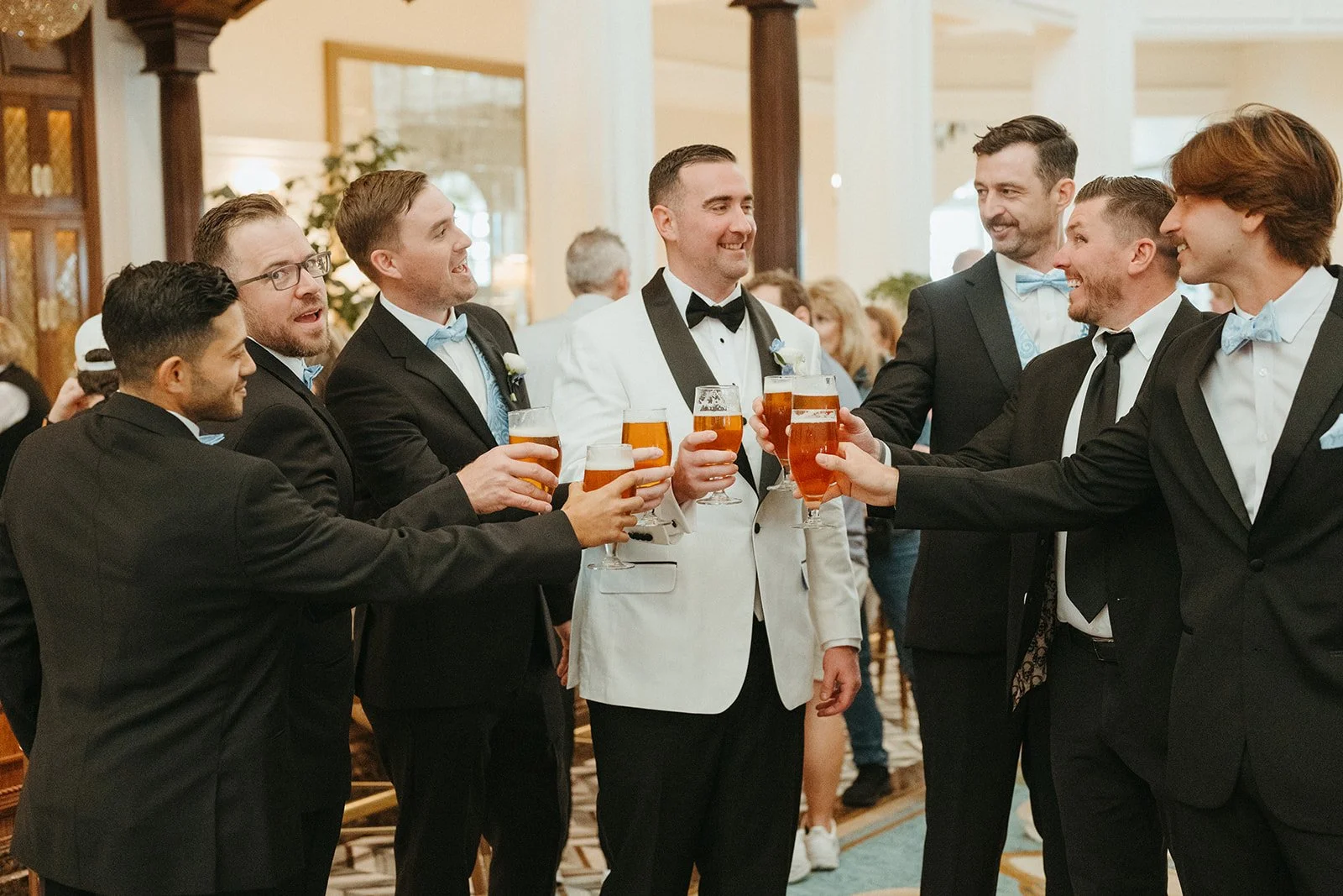 Group of men in tuxedos and one in a white tuxedo jacket, celebrating with drinks in a formal setting.