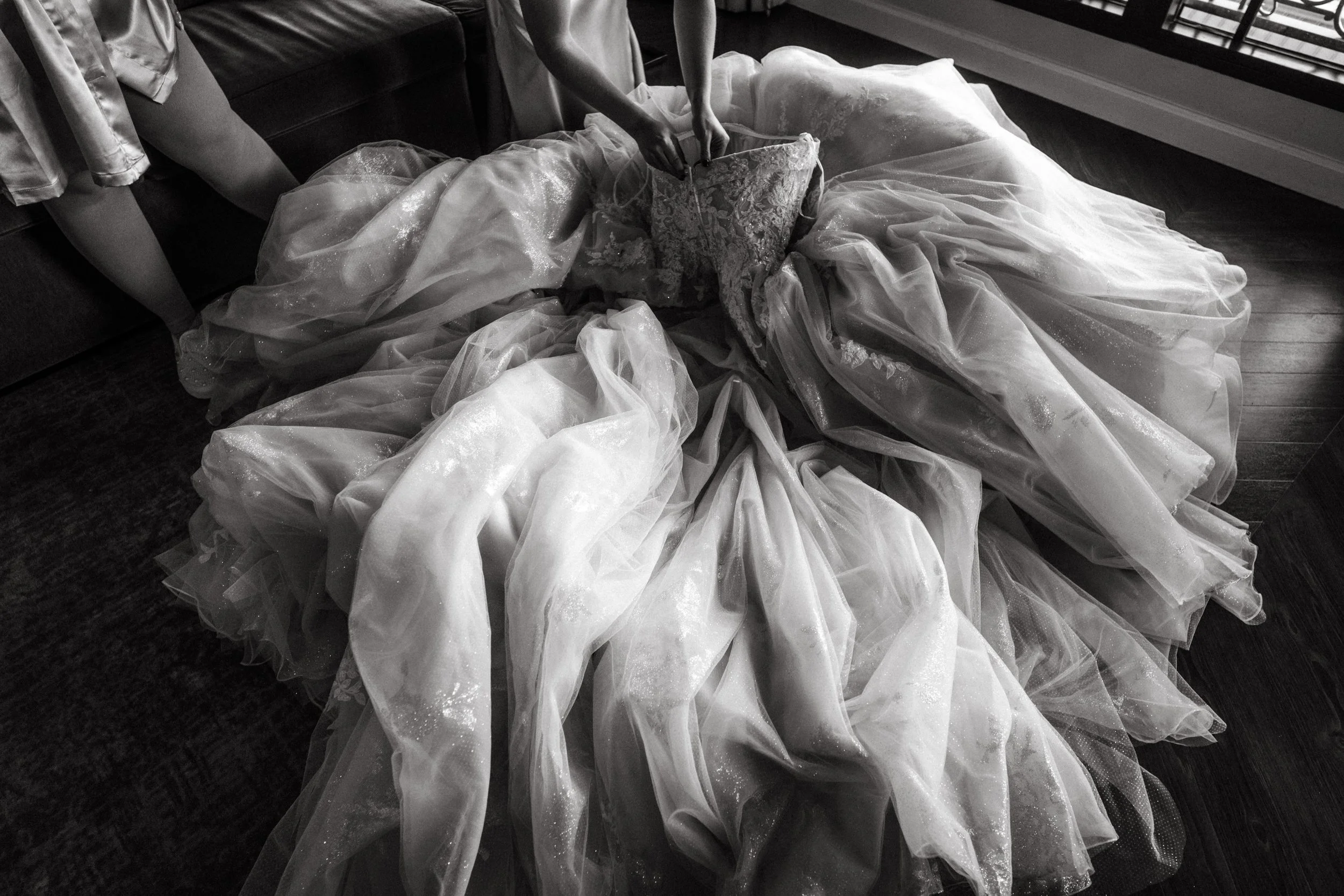A person in a wedding dress is kneeling on the floor, adjusting or arranging the gown's train, which is spread out in a circular fashion on a wooden floor. The dress is ornate with lace and tulle. There is a black sofa and a person wearing shorts par