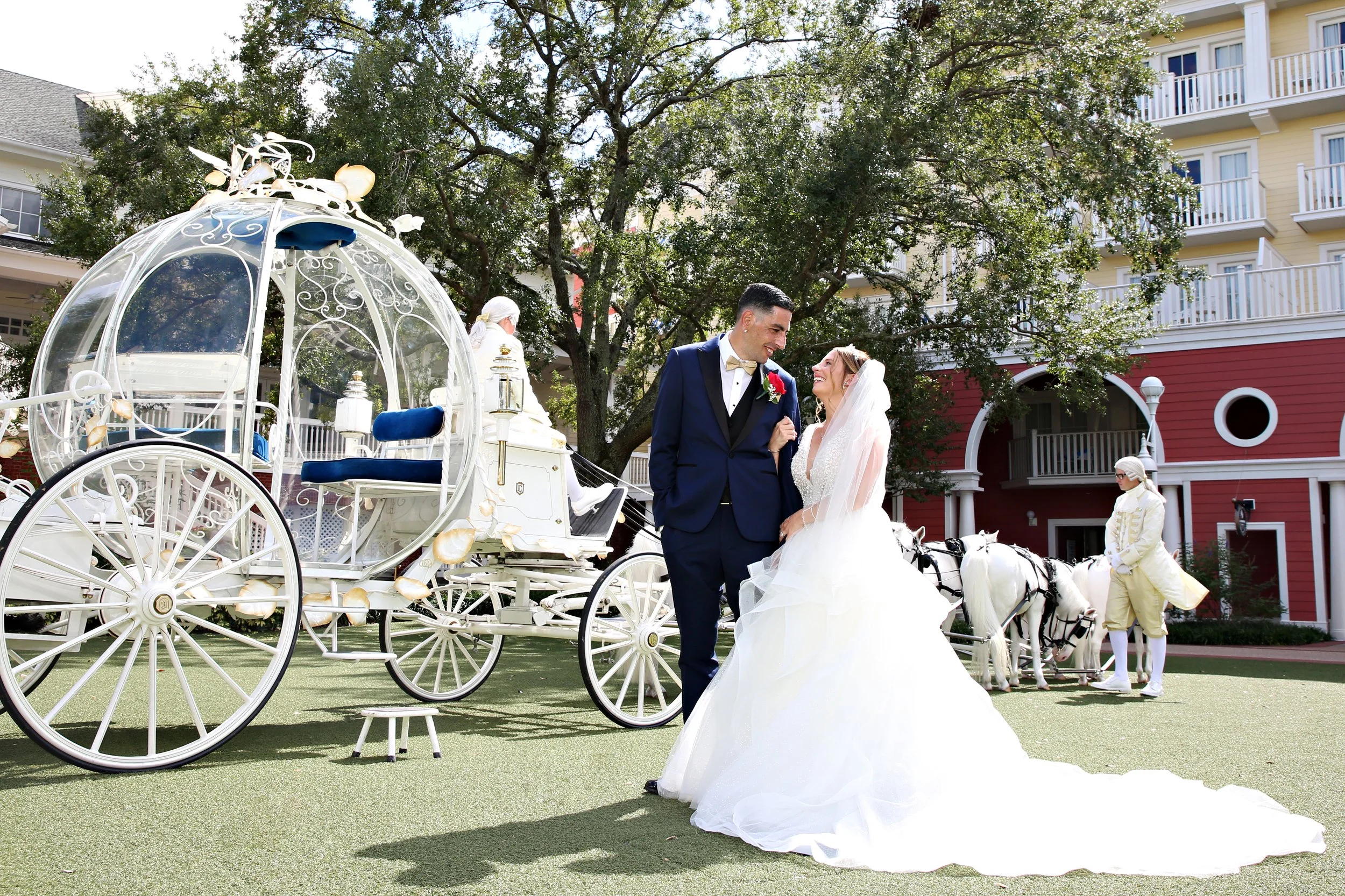 Bride and groom in wedding attire smiling and holding hands near a white carriage with blue seats, two knotted white horses, and stagecoaches in the background with staff members in vintage clothing, set outdoors with trees and buildings.