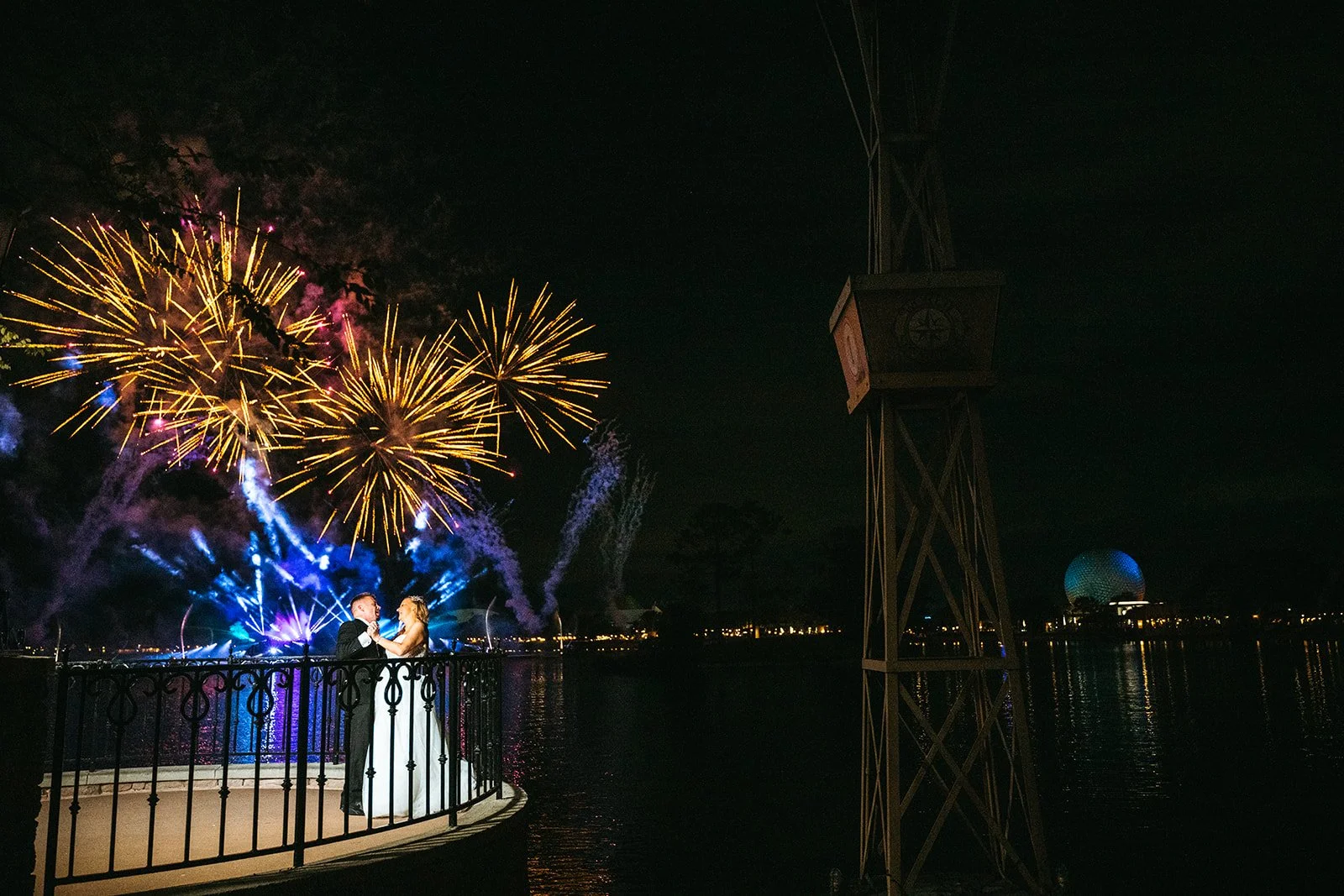 A bride and groom dancing on a small, fenced platform by a body of water during a fireworks display at night.
