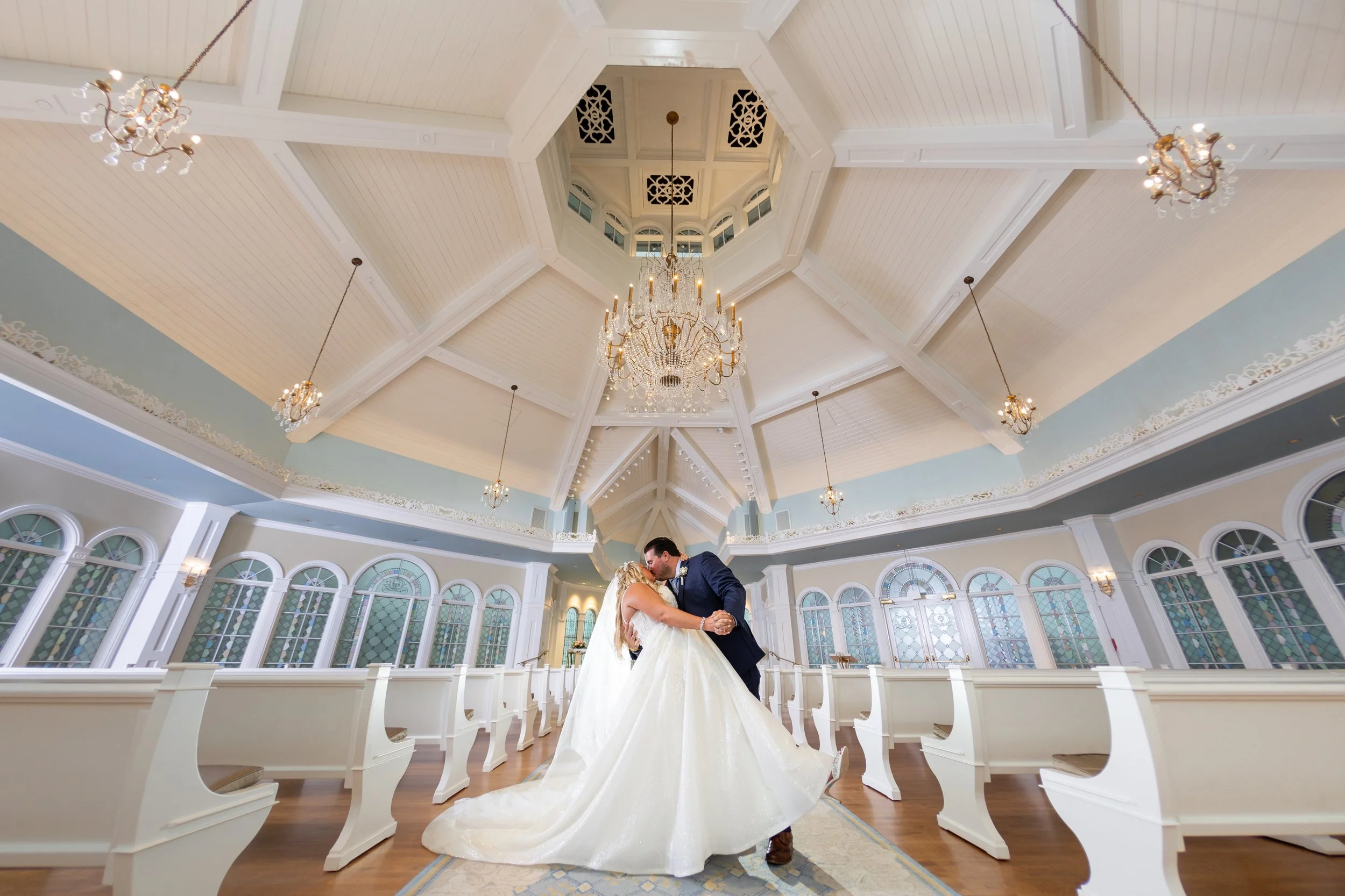 A bride and groom share a kiss in a bright, elegant wedding chapel with high vaulted ceilings, chandeliers, and stained glass windows.