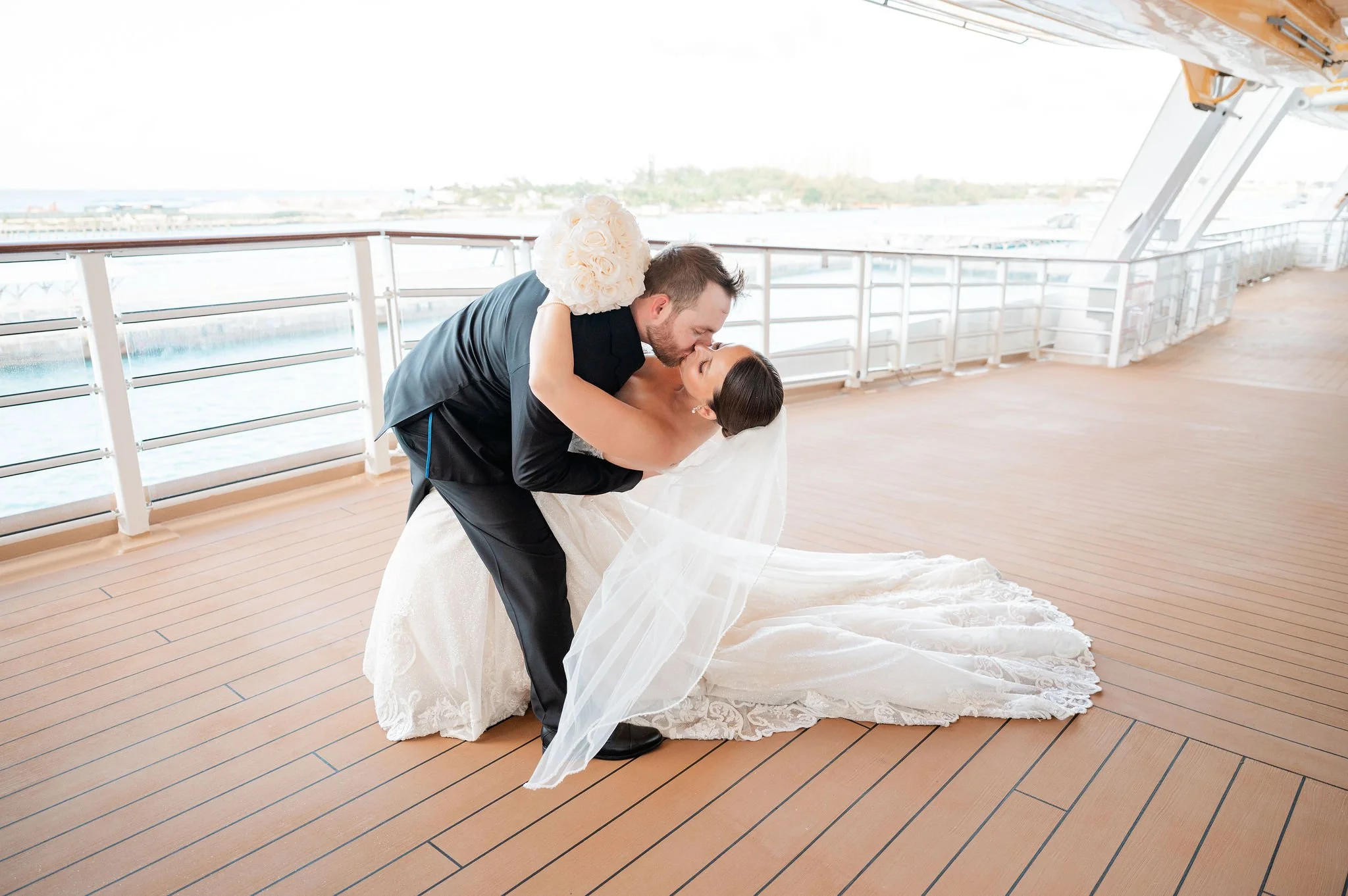 A newlywed couple sharing a kiss on a scenic ship deck, with the groom dipping the bride, dressed in a white wedding gown, holding a bouquet of white roses.
