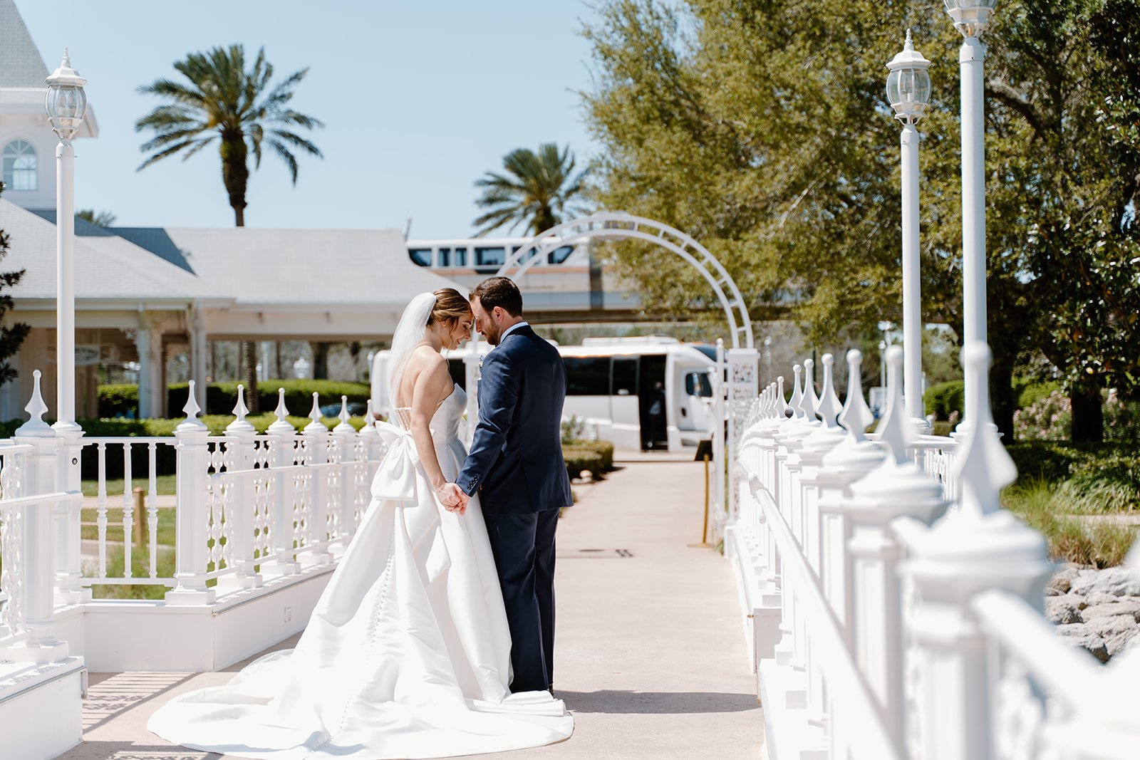 A bride and groom standing close with foreheads touching, holding hands outdoors on a sunny day, with white fencing and trees in the background.