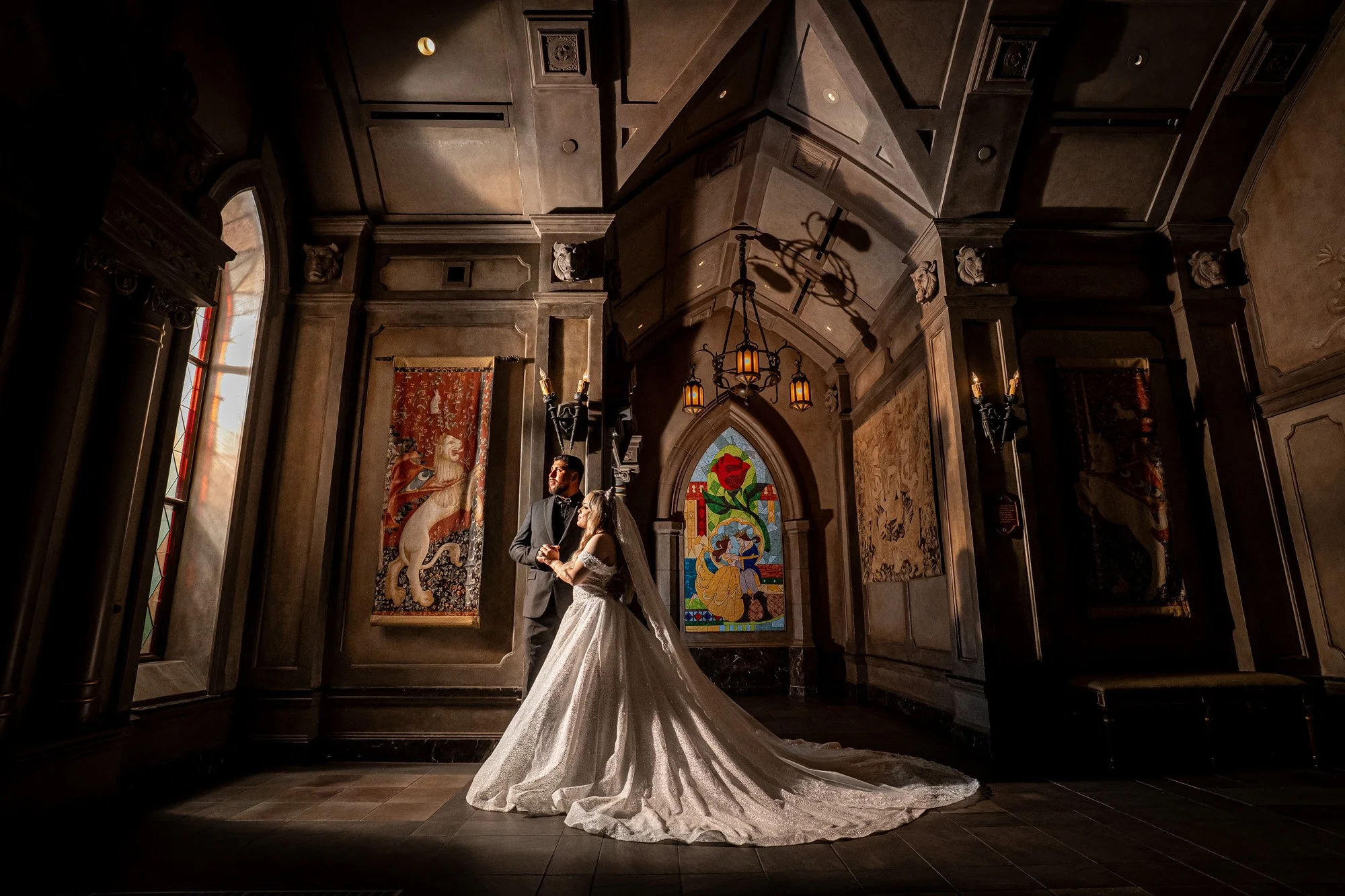 A bride and groom stand together in a grand, Gothic-style room with stained glass windows, ornate wall tapestry, and warm lighting.