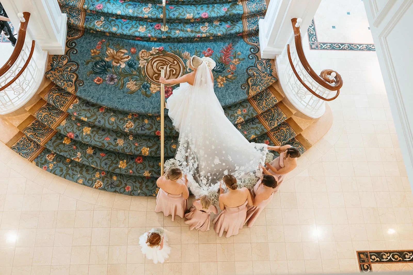 A bride in a white wedding gown standing on a grand staircase, surrounded by six bridesmaids in blush dresses, holding bouquets of white flowers, in a luxurious interior with ornate decor and a patterned carpet.