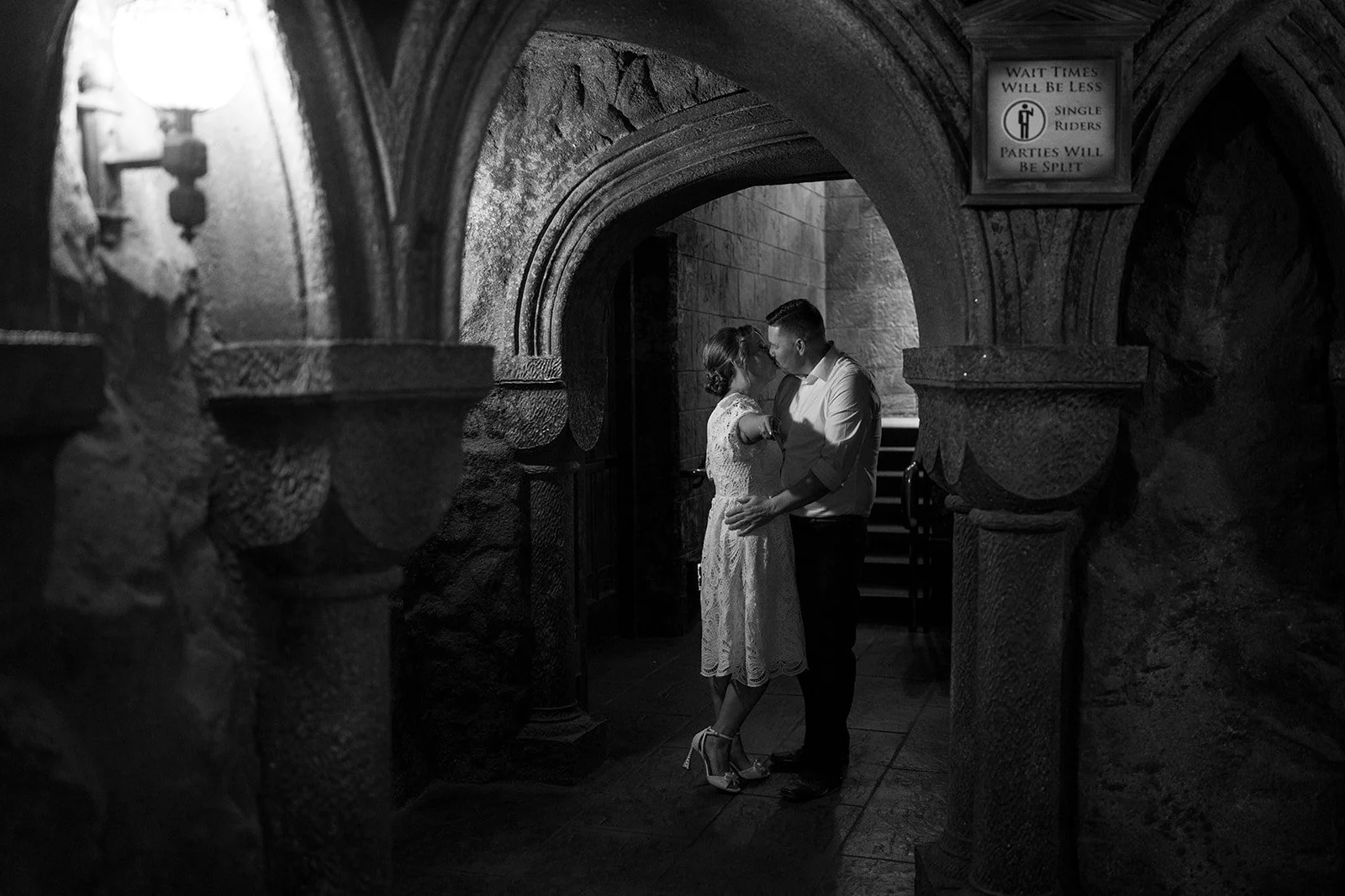 A couple sharing a kiss in a dimly lit, stone-archway hallway, with a sign above them indicating wait times and ride types.