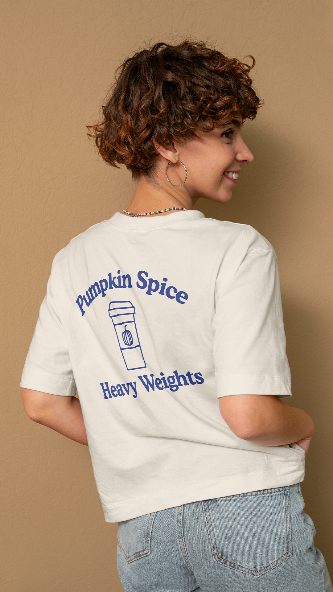 A person with curly brown hair wearing a white t-shirt with a graphic that says "Pumpkin Spice Heavy Weights" and a drawing of a to-go coffee cup, standing against a beige wall.