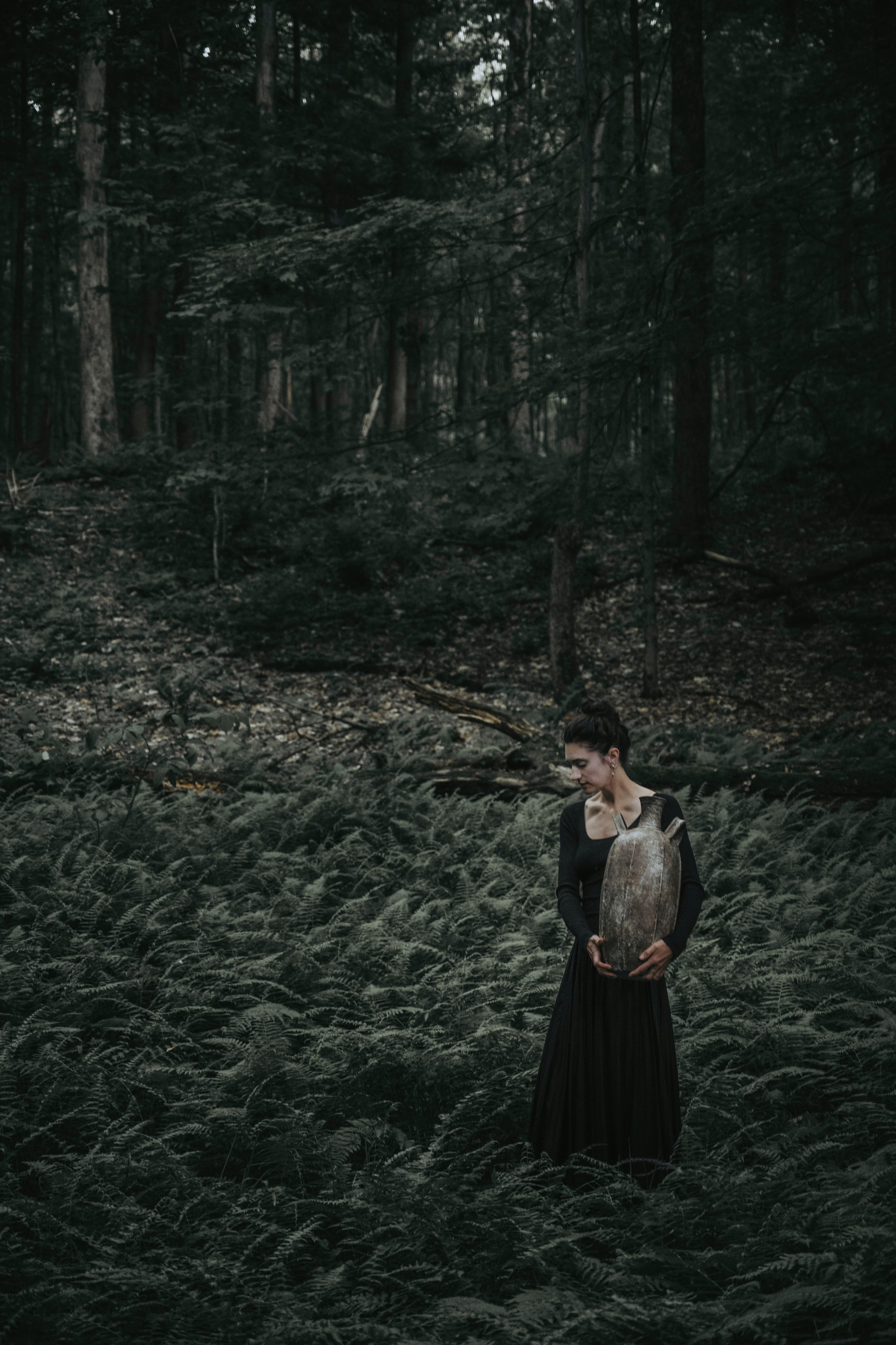 A woman in a black dress and long skirt stands in a dense forest, holding a large, ceramic vessel against her body. The forest is dark and full of green ferns and tall trees.