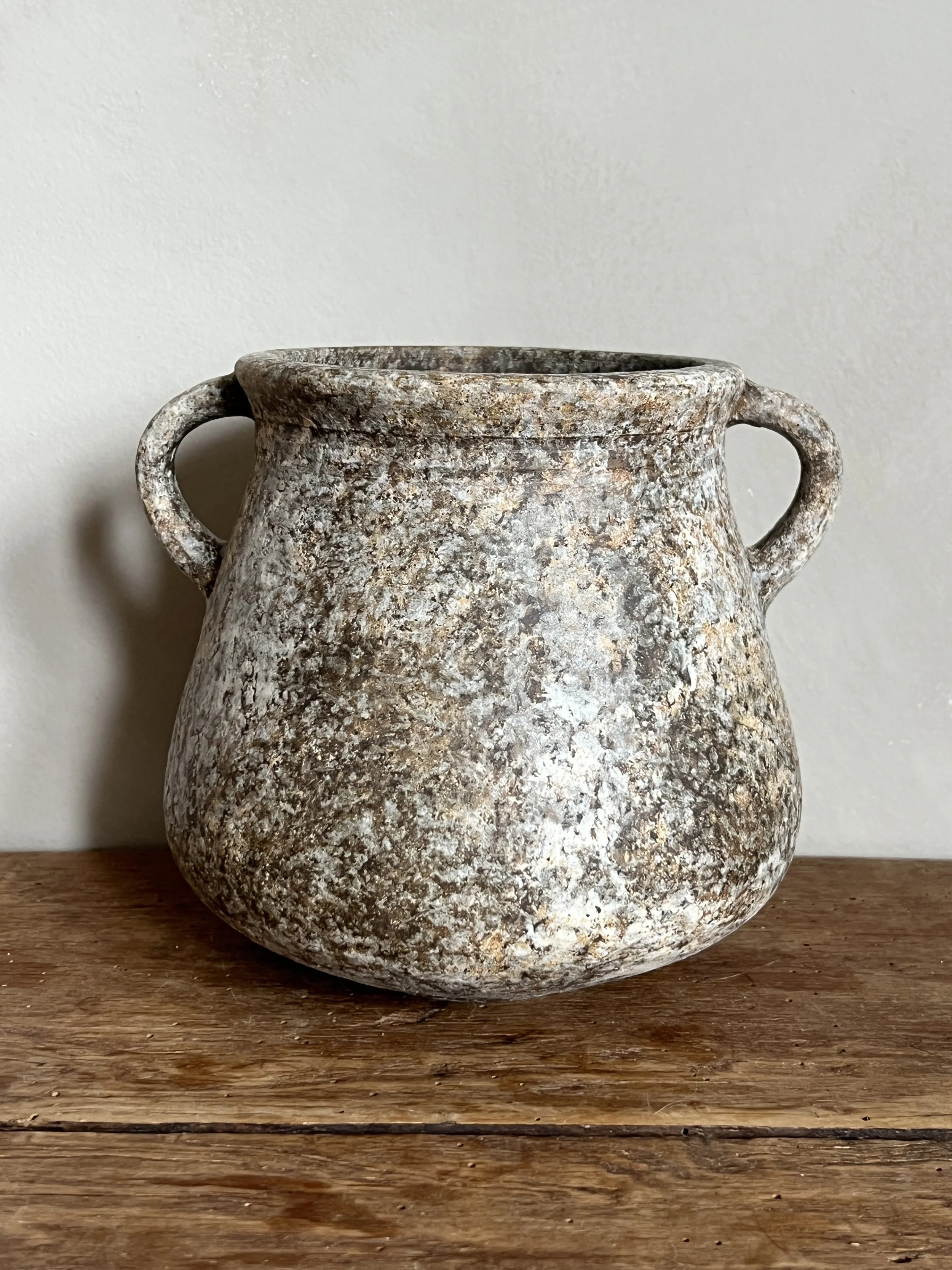A weathered, antique stoneware jar with two small handles, sitting on a wooden surface against a plain wall.
