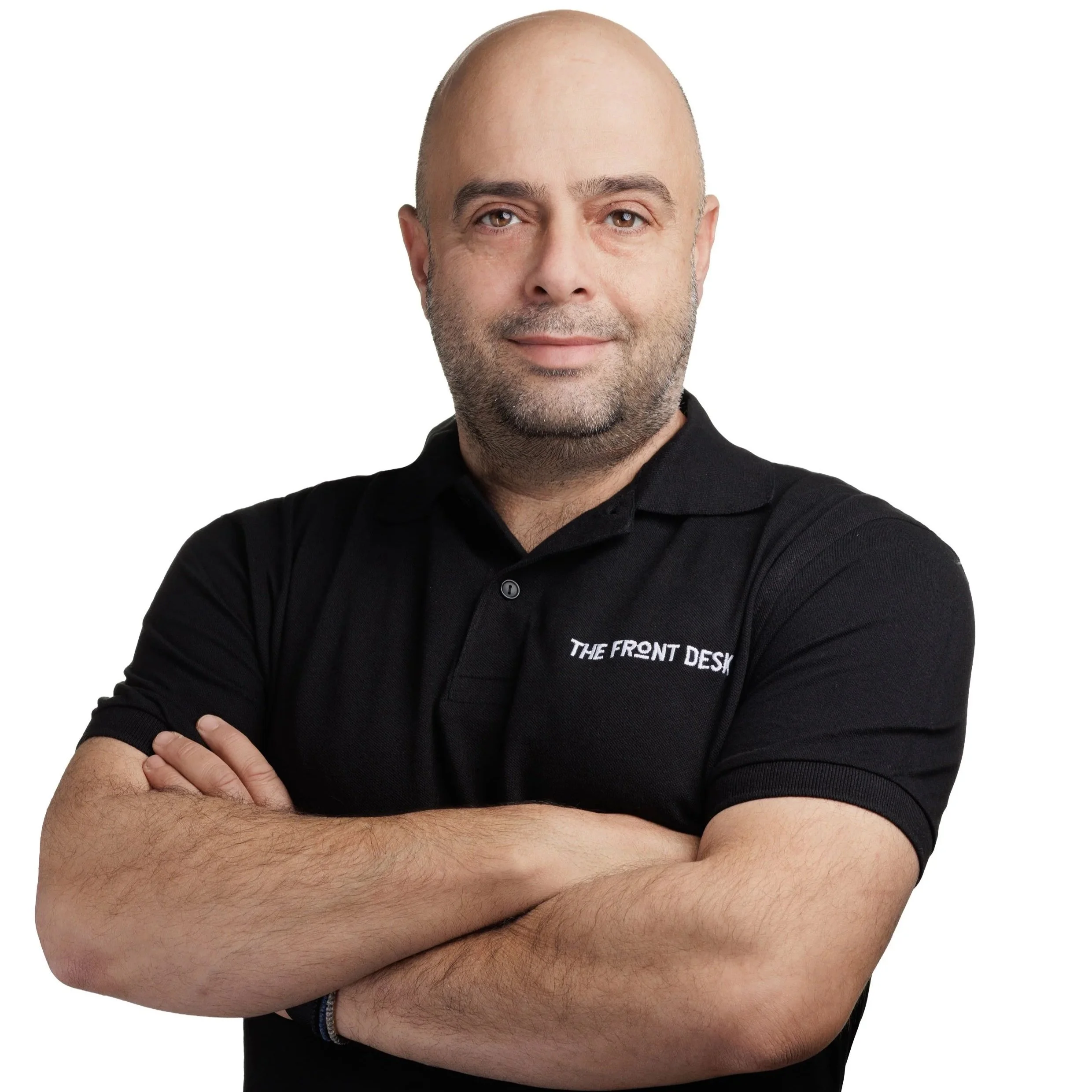 Man with a shaved head and beard crossing arms, wearing a black polo shirt with 'THE FRONT DESK' embroidered.