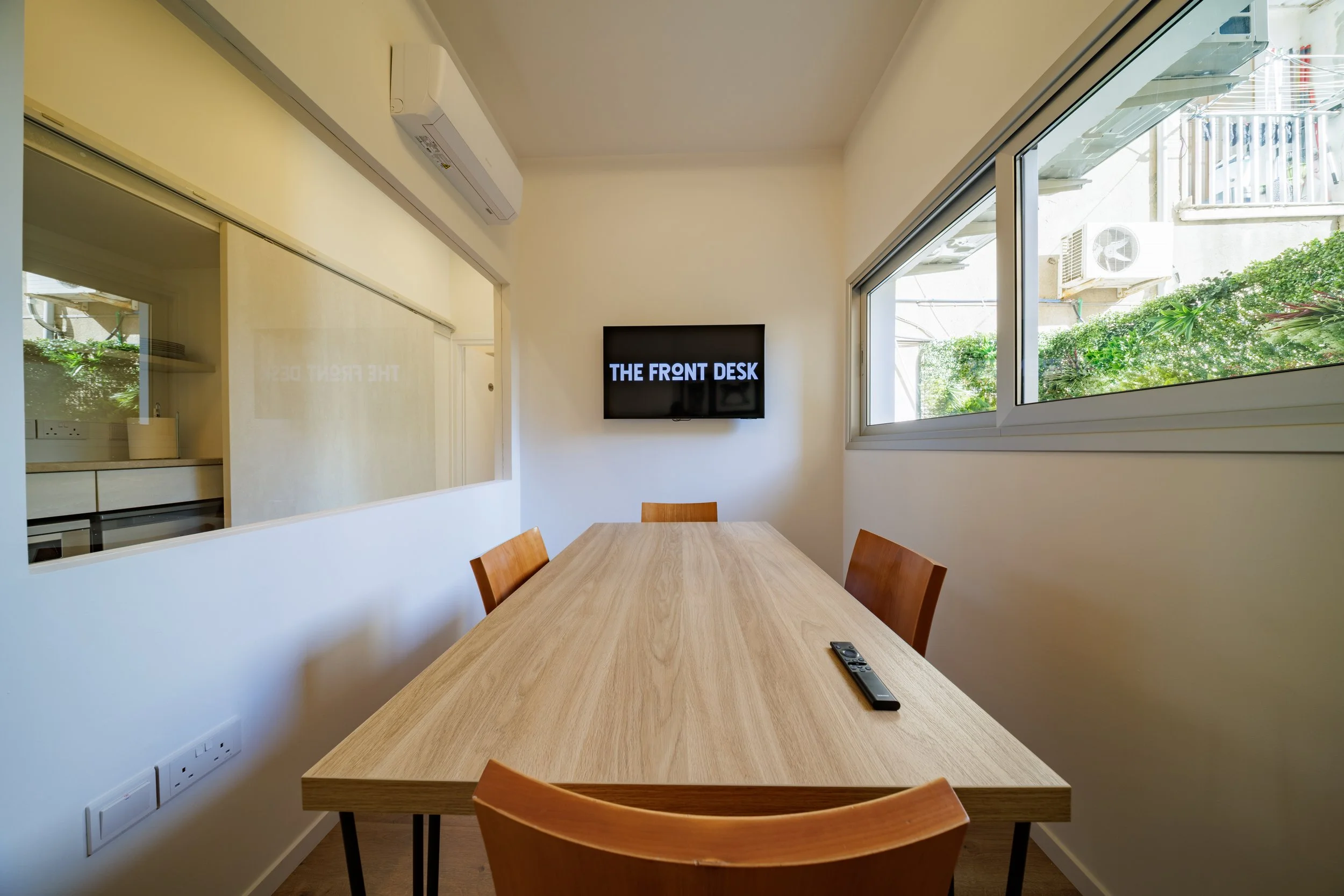 Meeting room with a wooden table, four chairs, a wall-mounted TV displaying 'THE FRONT DESK,' large windows with greenery outside, and a small remote control on the table.