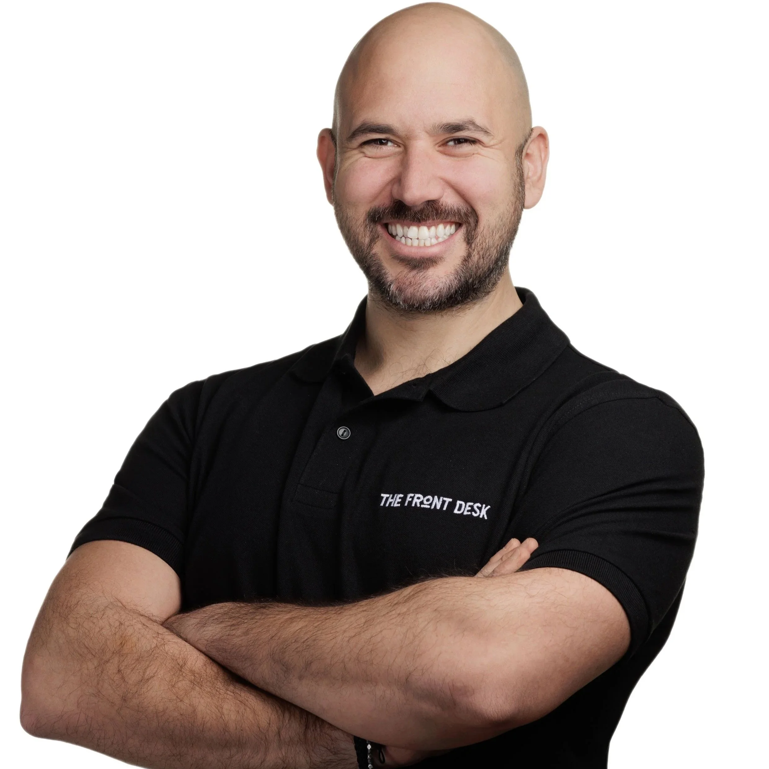 A smiling man with a beard and bald head, wearing a black polo shirt with white embroidery that says 'THE FRONT DESK'. He has arms crossed and is standing against a white background.