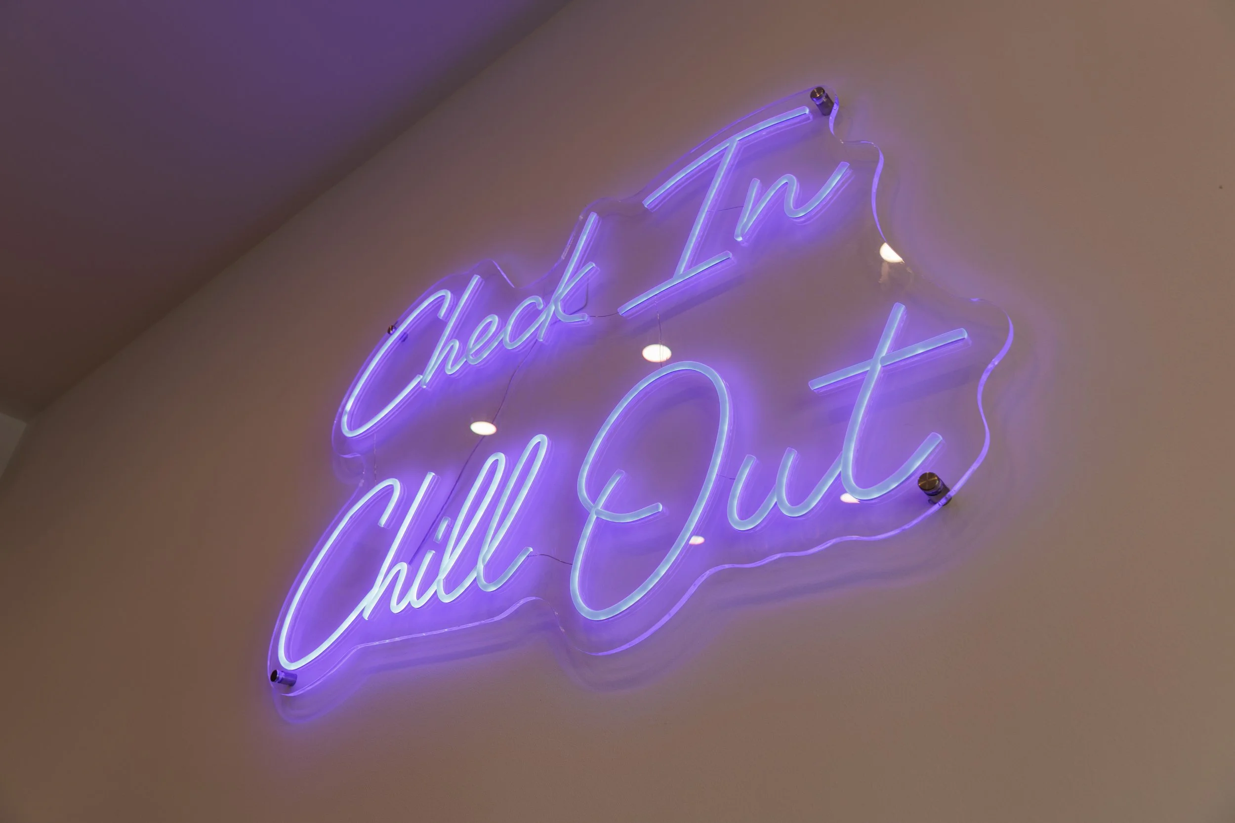 A purple neon sign on a wall that says, "Check In, Chill Out" in cursive script.