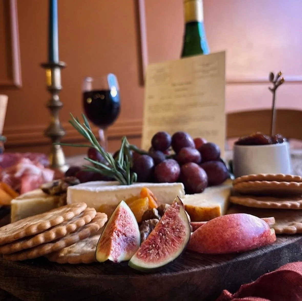 A cheese and fruit platter with figs, grapes, crackers, and cheese on a wooden board, with a glass of red wine and a candle in the background.