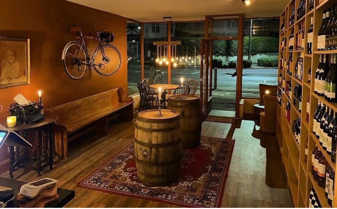 Interior of a cozy wine shop with wooden barrels as tables, a vintage bicycle mounted on the wall, and shelves filled with wine bottles. There is a window and door open to the street outside, with warm lighting and a small seating area.