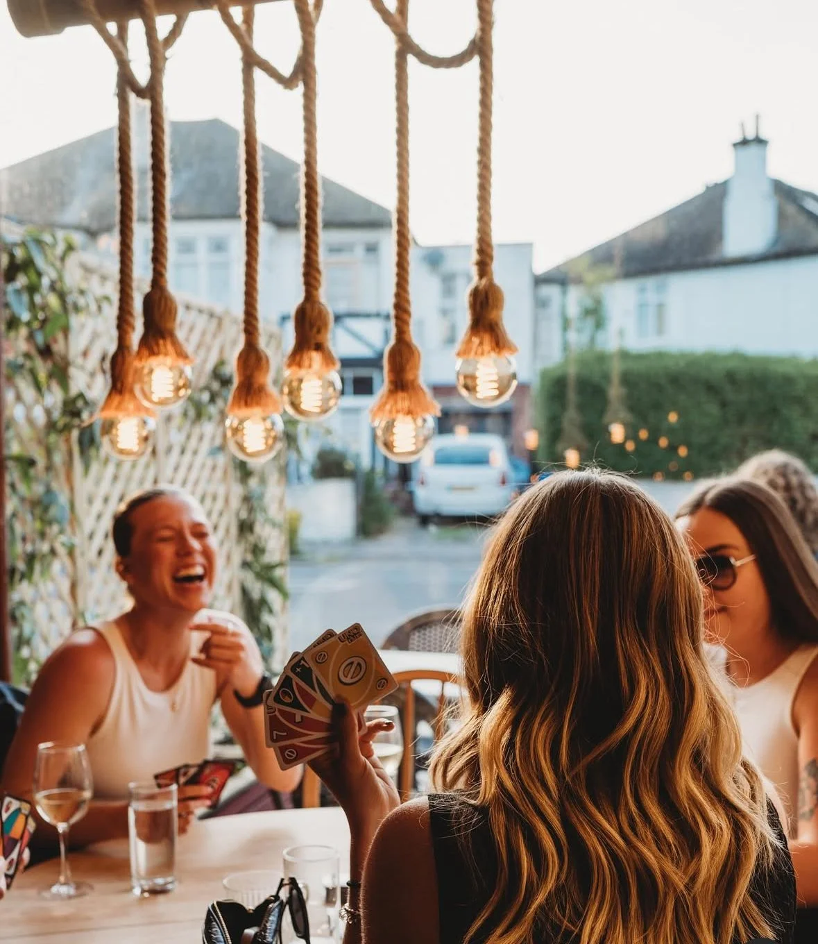 A group of women playing a card game at an outdoor gathering, with string lights hanging overhead and a residential street in the background.