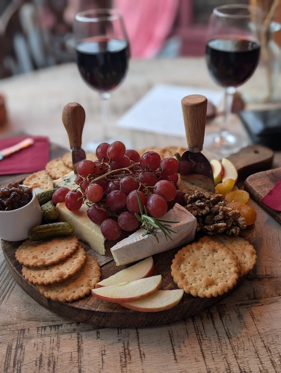 Cheese and fruit platter with grapes, apple slices, crackers, walnuts, and a small bowl of jam, served on a wooden board with two glasses of red wine in the background.