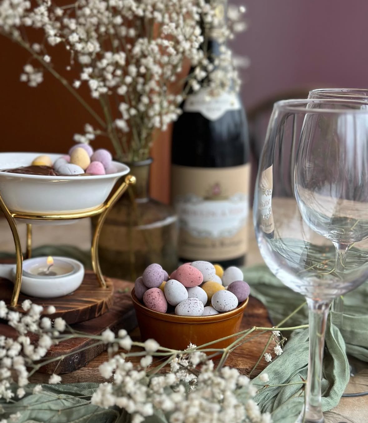 Table setting with pastel-colored candy eggs, empty wine glasses, a candle, a small bowl with chocolate, and decorative flowers in the background.