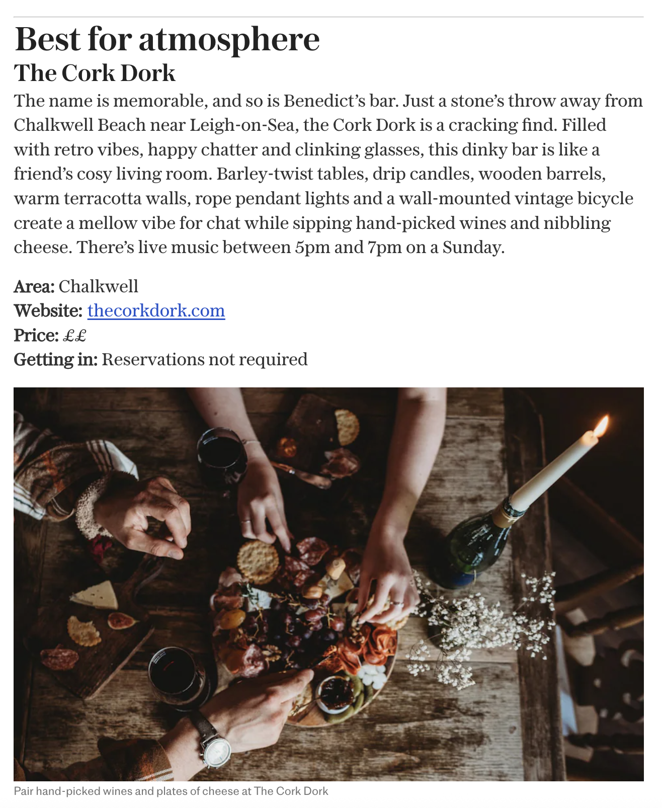 People reaching for cheese and wine on a wooden table with a candle, flowers, and snacks.