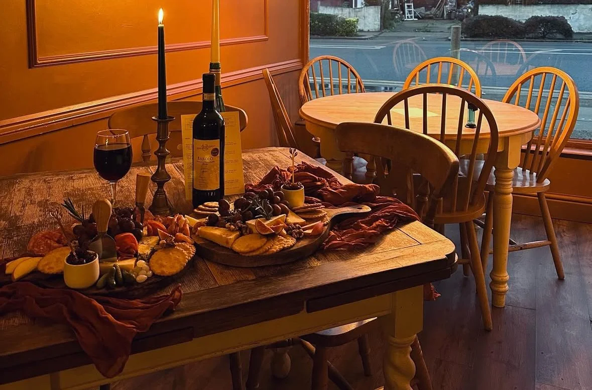 A rustic wooden dining table set with a cheese and fruit platter, a bottle of red wine, a glass of wine, and candles in a cozy restaurant with wooden chairs and a large window in the background.