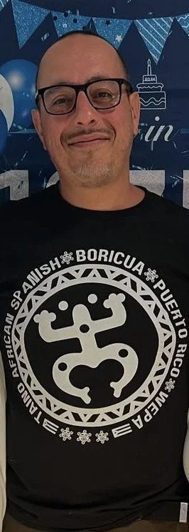 Man with glasses smiling, wearing a black T-shirt with a white circular logo featuring a gecko and text. Background has celebration decorations including balloons, banners, and a cake with a candle.