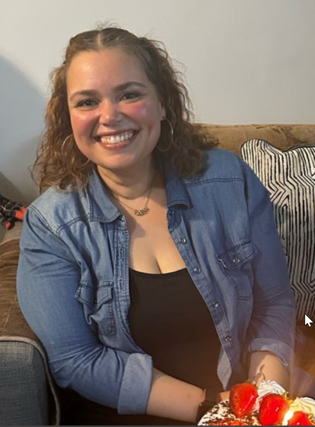 A young woman with curly hair and hoop earrings smiling, sitting on a brown couch with striped pillows, holding a birthday cake with strawberries.