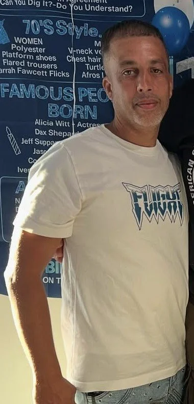 A man with short dark hair and light skin standing indoors near a blue poster and balloons, wearing a white T-shirt with a graphic logo and light-colored pants.