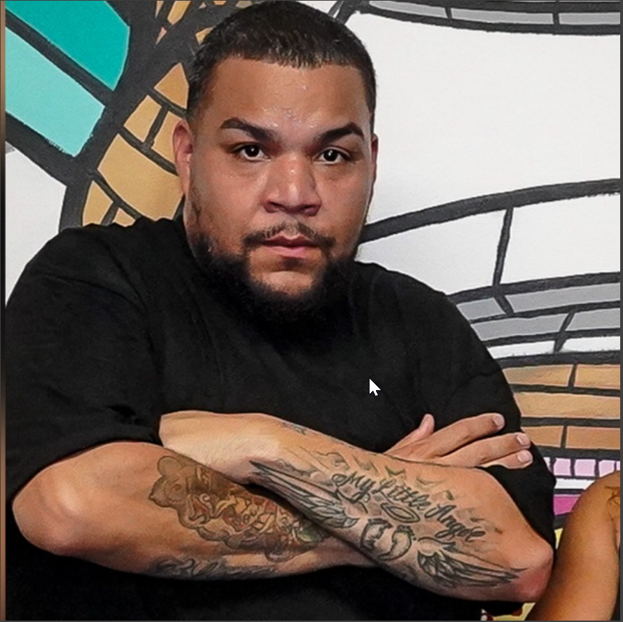 A man with short dark hair, a beard, and tattoos on his arms, wearing a black shirt, sitting with arms crossed in front of a colorful, abstract mural.