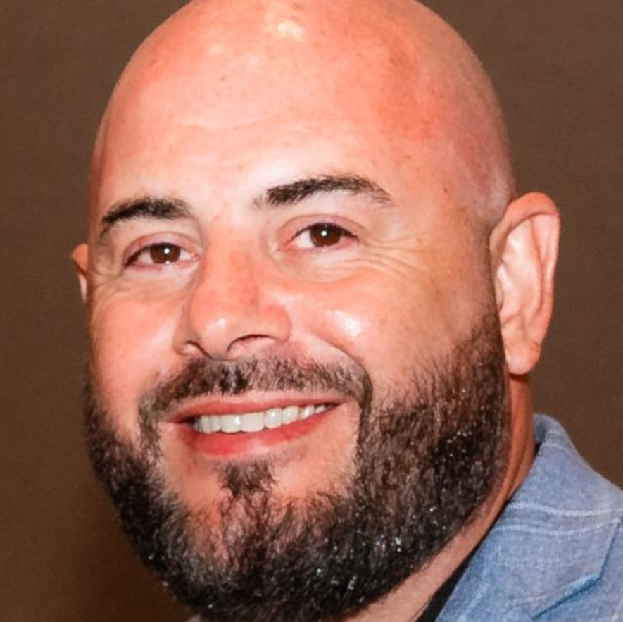 Close-up of a smiling bald man with a beard, wearing a gray suit jacket.