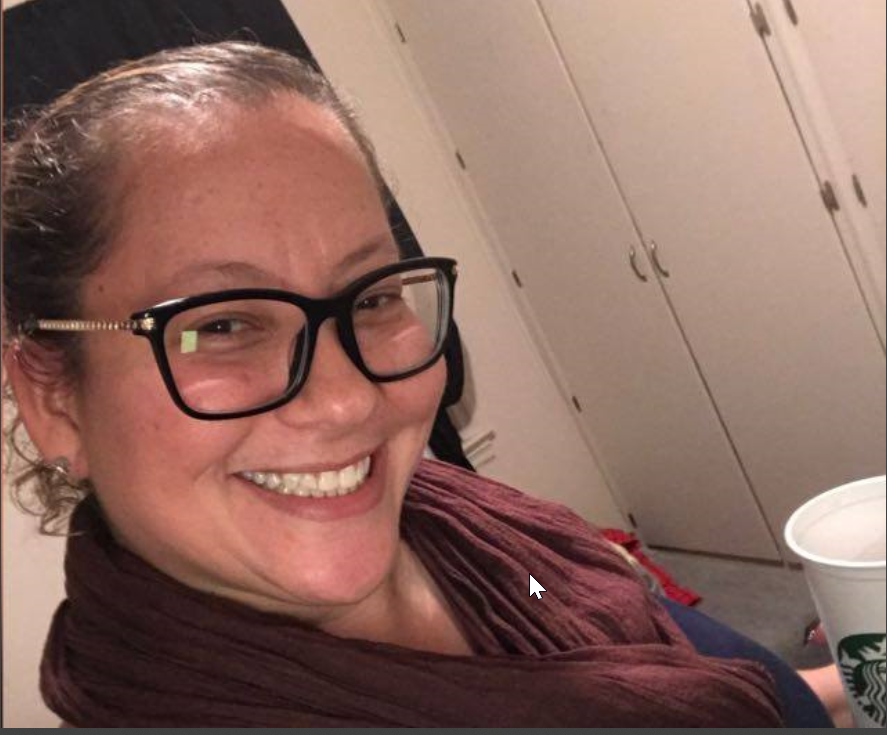 A woman with curly hair pulled back, wearing glasses and a maroon scarf, smiling and holding a Starbucks cup.