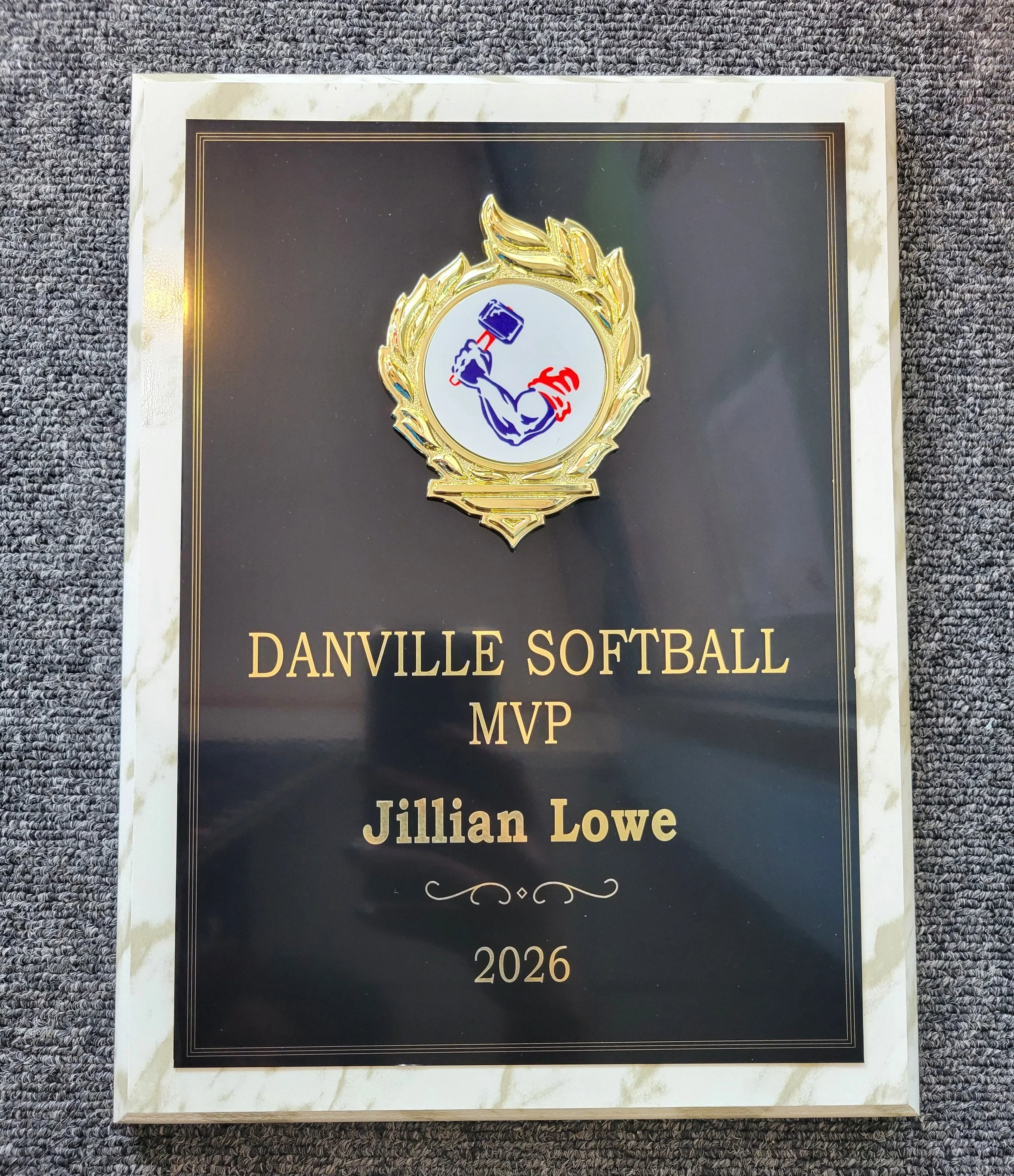 Award medal with a muscular arm holding a hammer, surrounded by a gold laurel wreath, on a black and white marble plaque with gold text reading "Danville Softball MVP Jillian Lowe 2026."