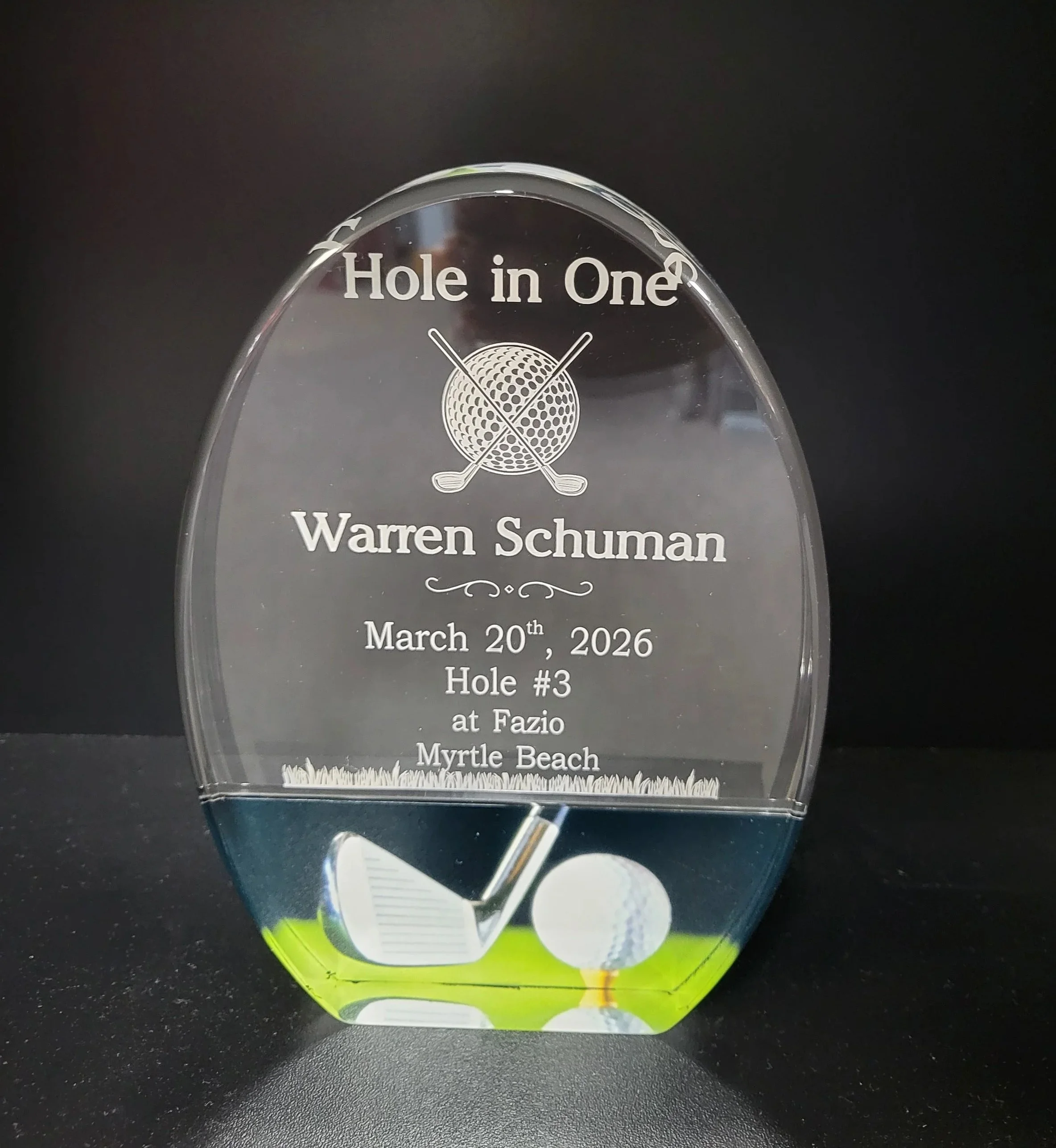 A glass award with engraved text including the words "Hole in One," the name Warren Schuman, and the date March 20th, 2026. The award has a golf ball and two crossed golf clubs engraved on it and is placed on a black surface.