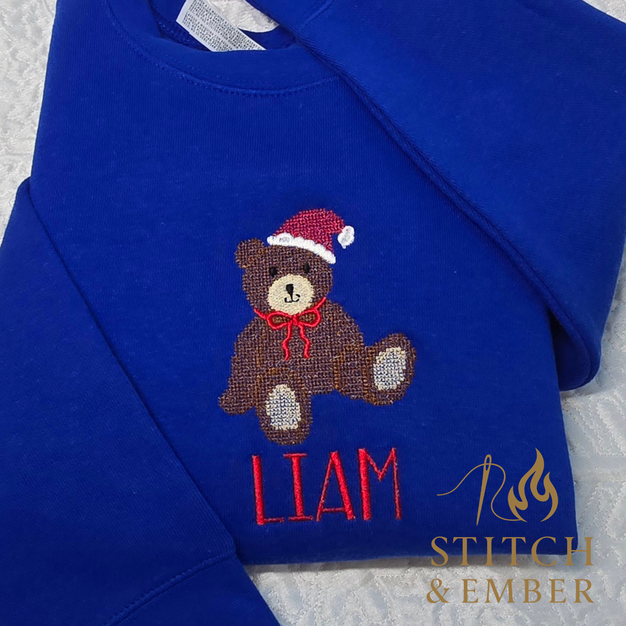 Blue sweatshirt with an embroidered teddy bear wearing a Santa hat and red bow, and the name Liam stitched in red below.
