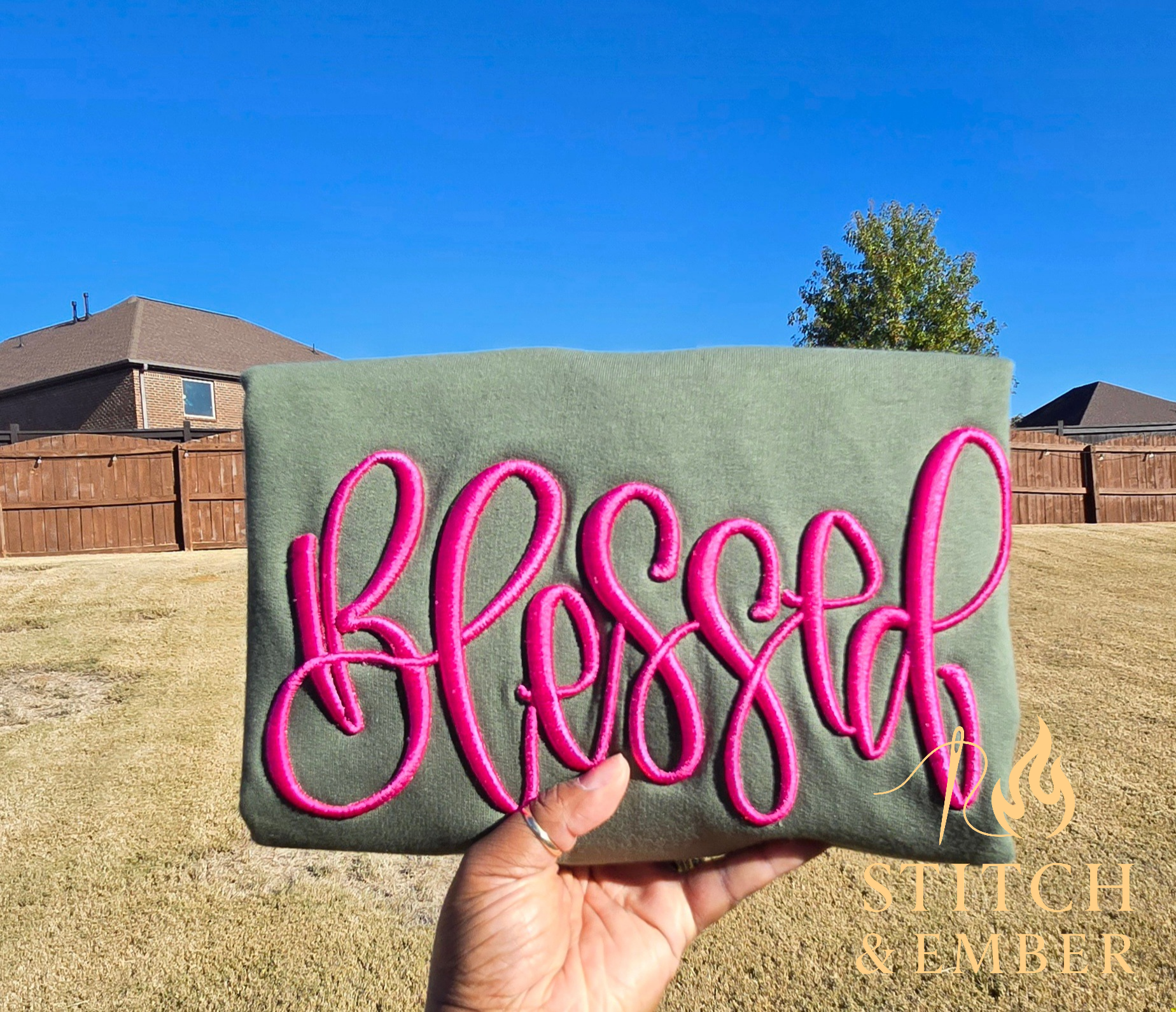Hand holding a green folded sweatshirt with pink embroidered word "Blessed" in a backyard with a brown fence, a single tree, and houses under a clear blue sky.