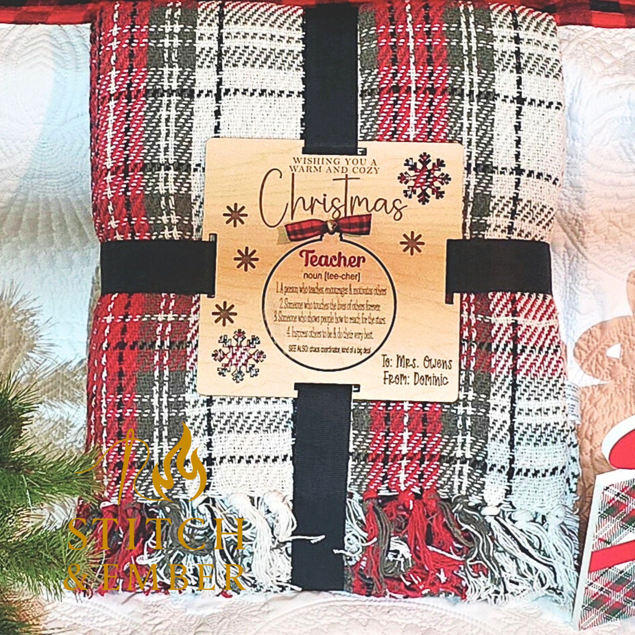 A red, black, and white plaid throw blanket with black straps and a engraved blanket tag attached, featuring holiday greetings and a rhyme about a teacher, with colorful snowflake illustrations and a green pine branch in the foreground.